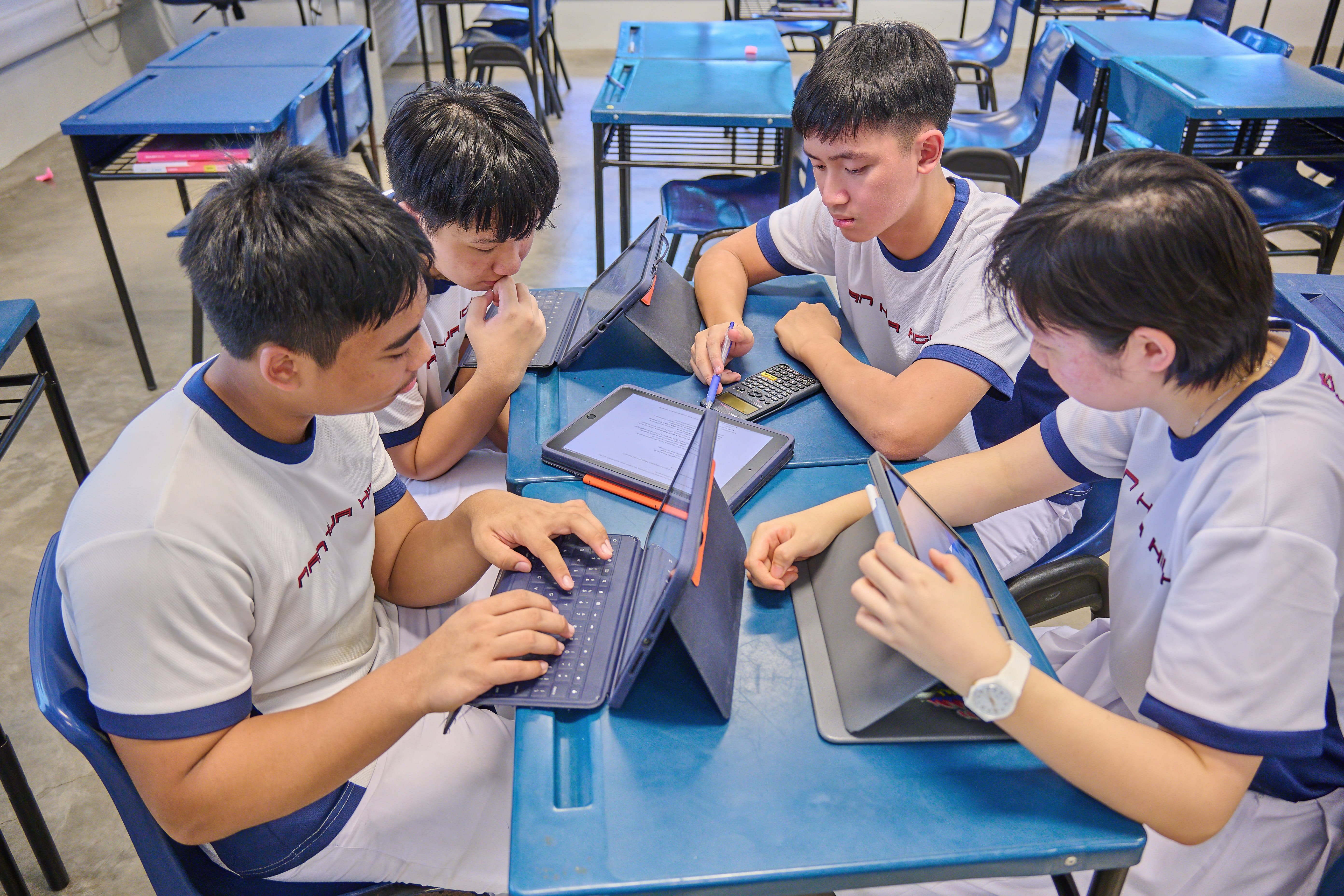 Four students in white uniforms sit around a blue desk, working together on tablets and a keyboard in a classroom.