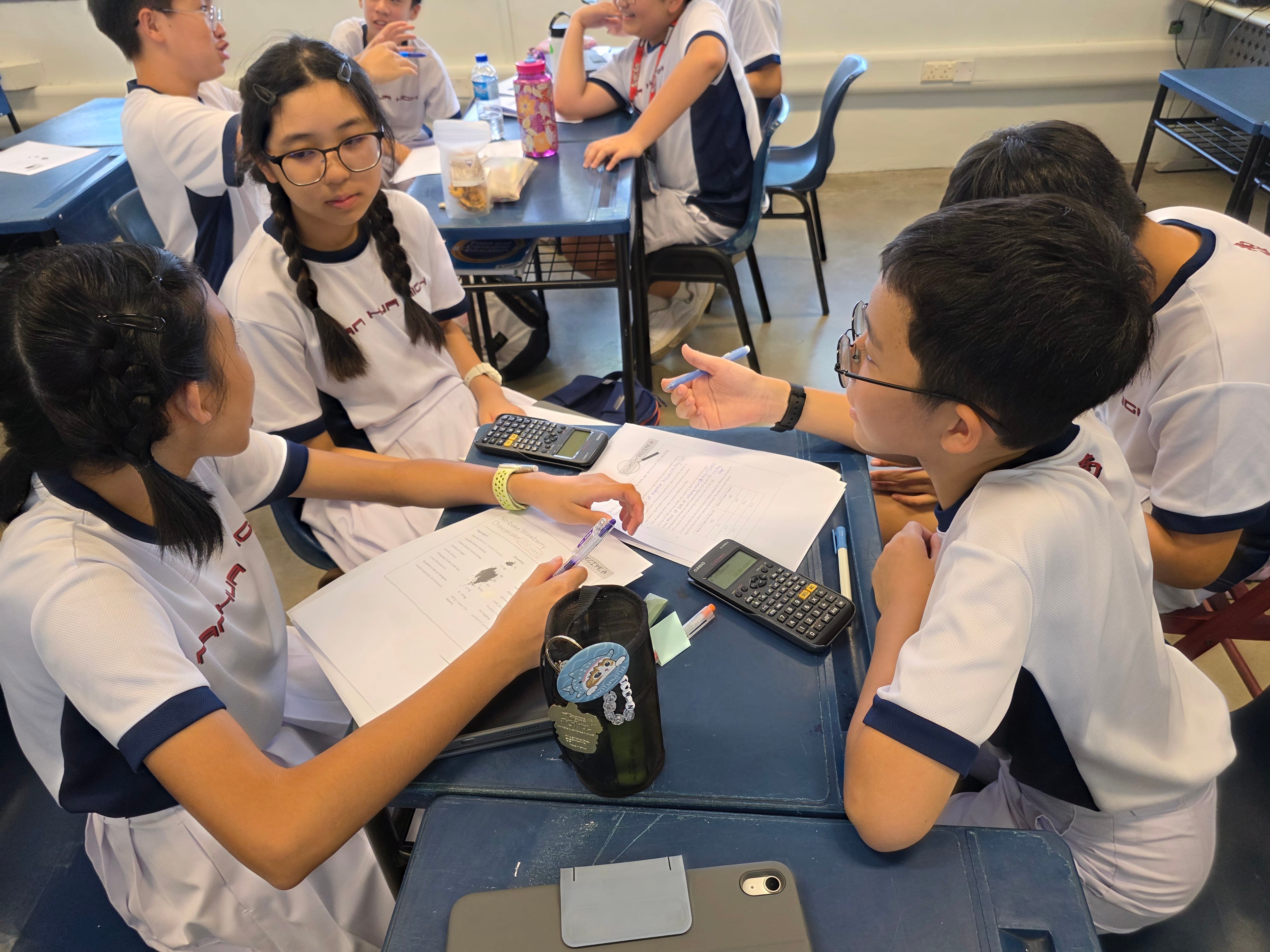 Four students in white uniforms discuss worksheets and calculators at a desk in a classroom.