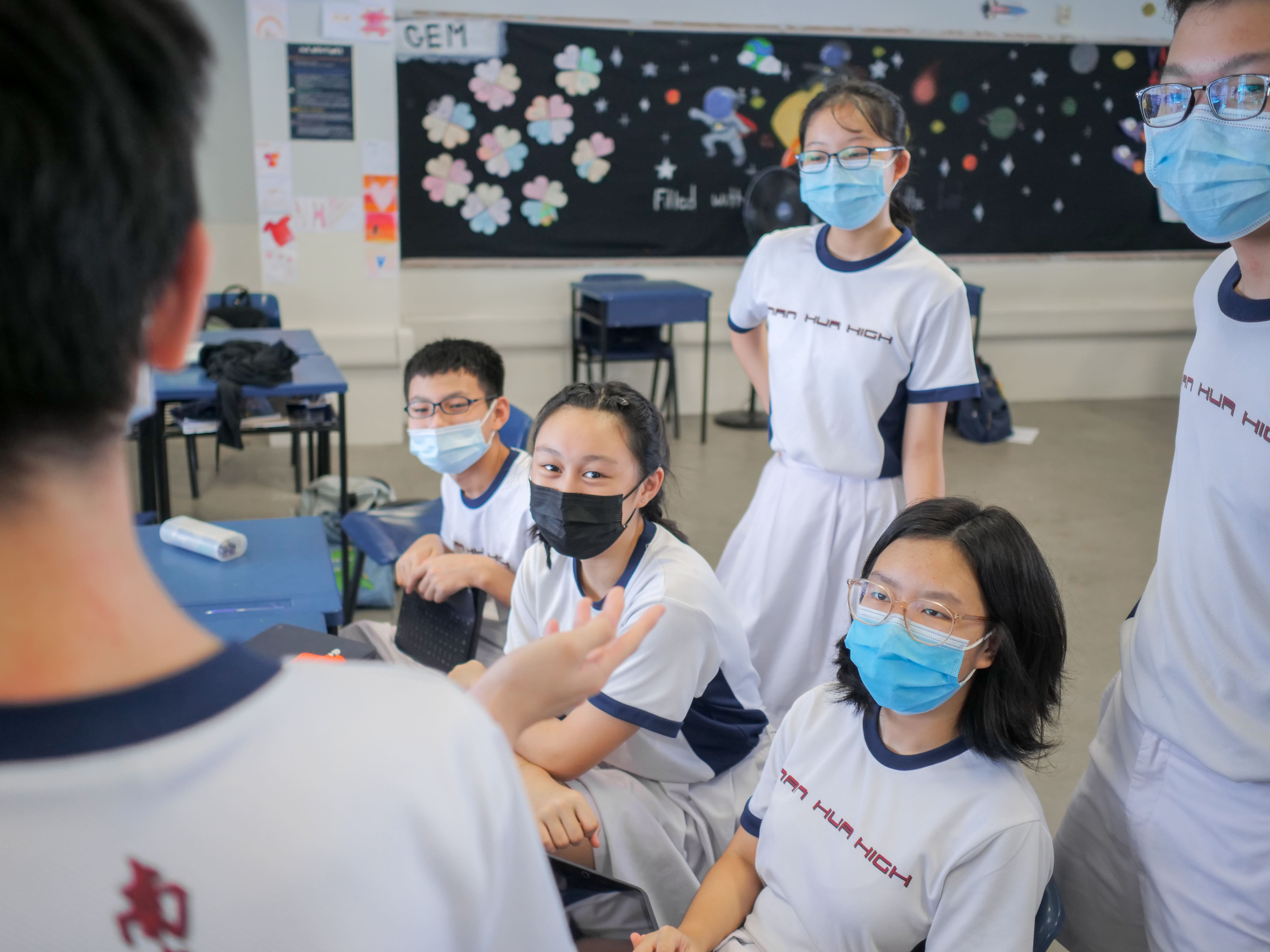 Several students in Nan Hua High uniforms and face masks gather in a decorated classroom, listening to a peer.