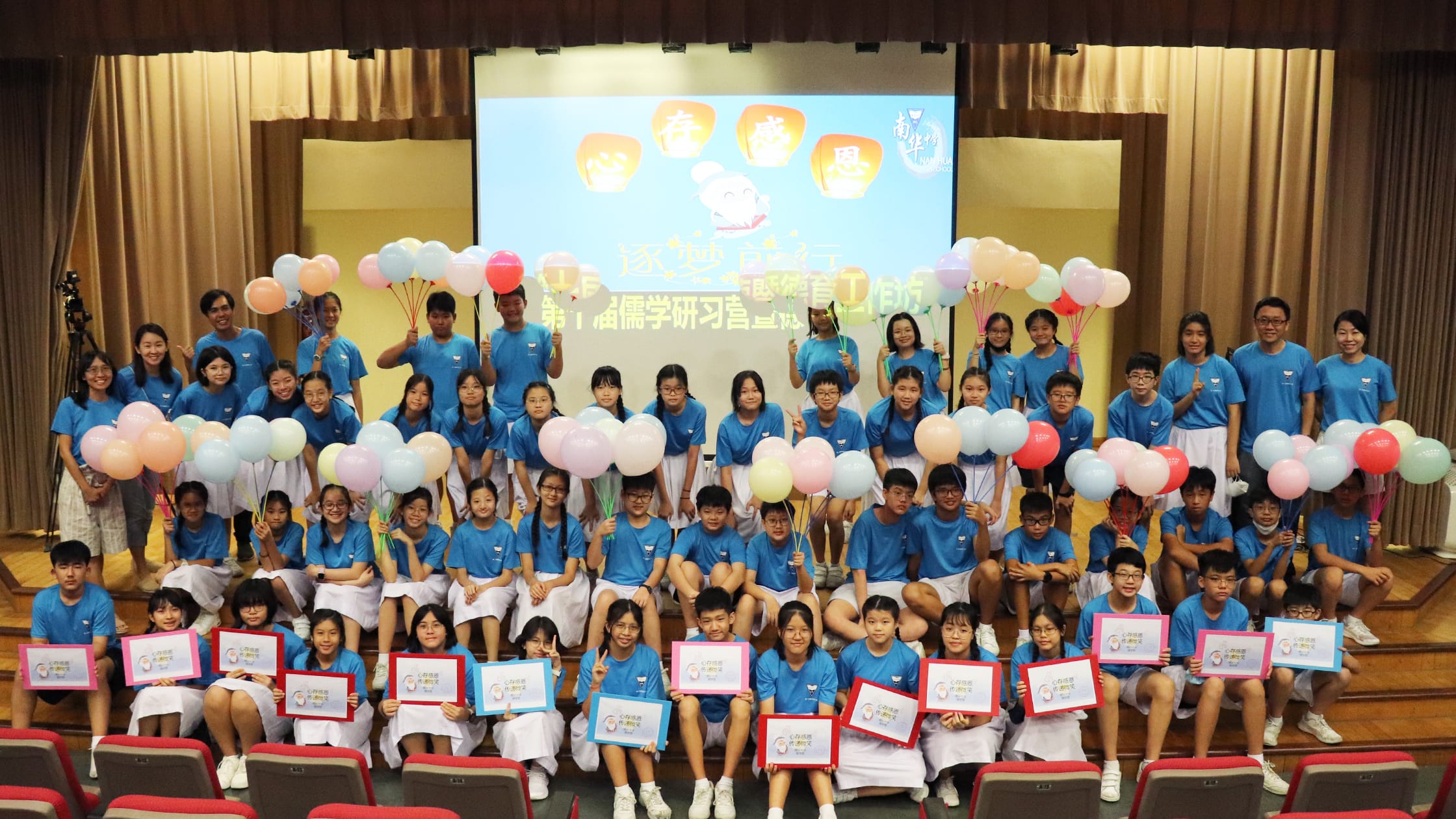 Large group of students in blue shirts pose on a stage holding colorful balloons and signs, with a Chinese banner behind them.