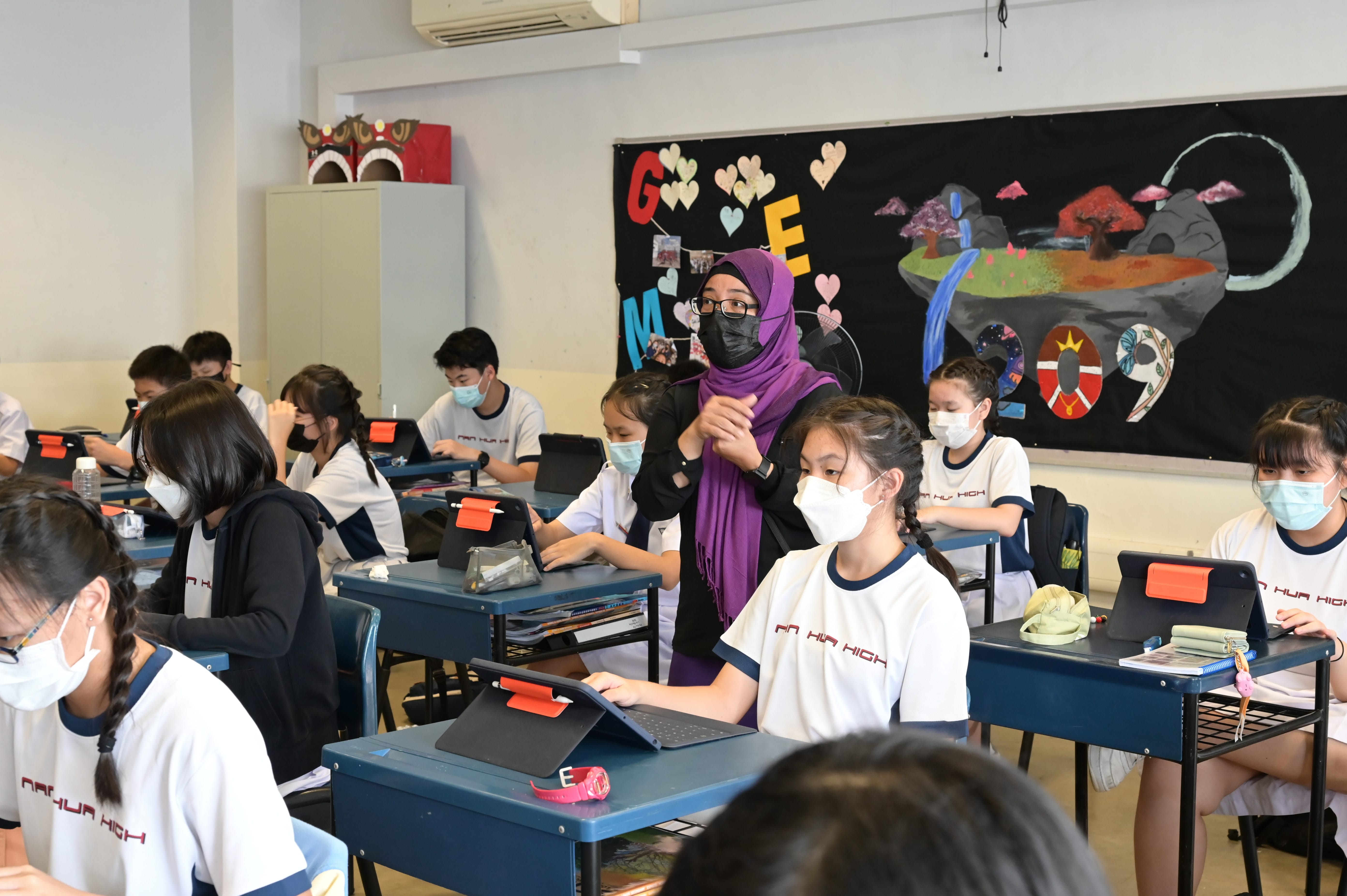 A teacher in a purple hijab and mask walks among masked students using tablets at desks in a decorated classroom.