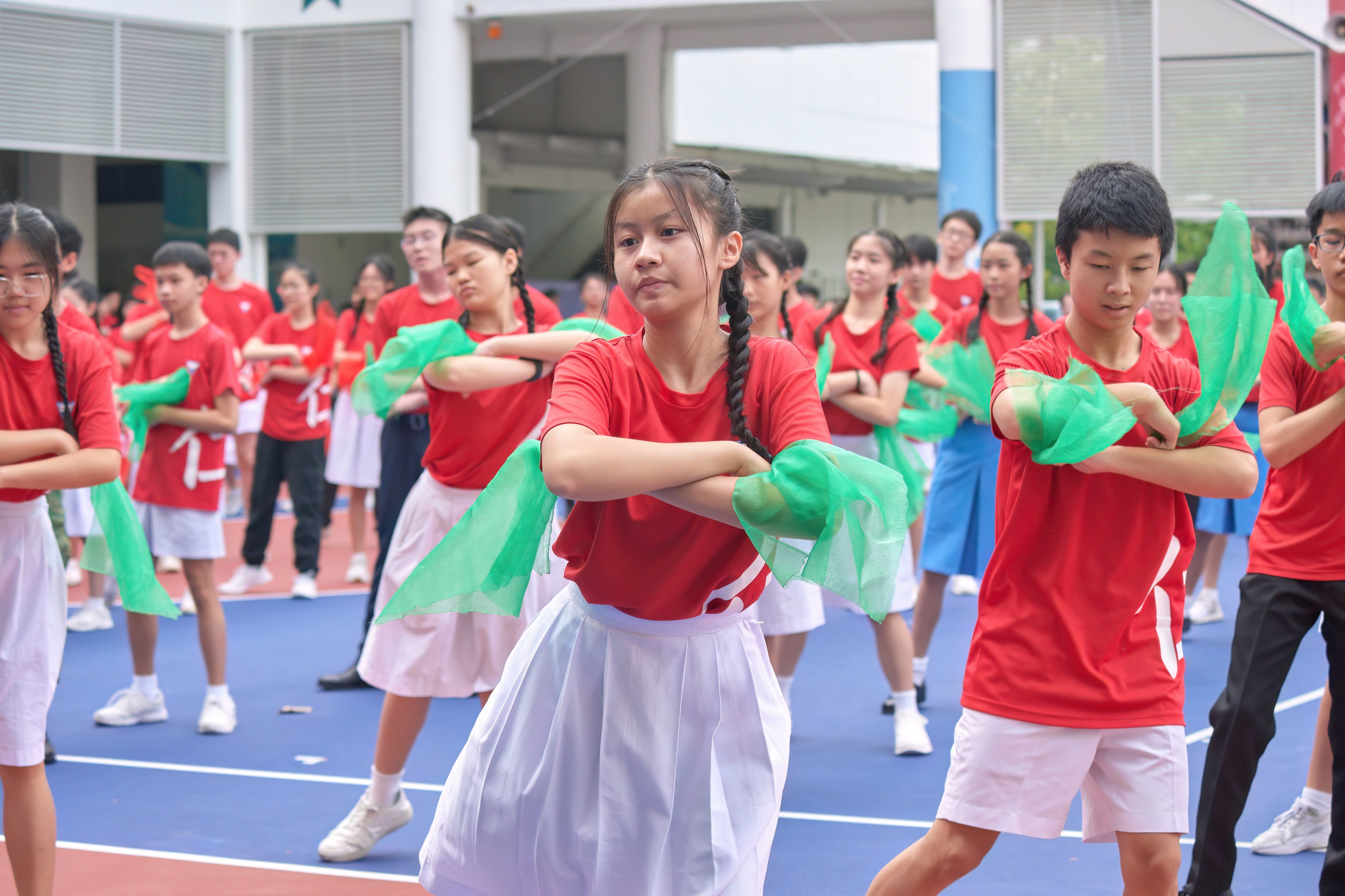 Students in red shirts dance with green scarves on a blue outdoor court during a group performance.