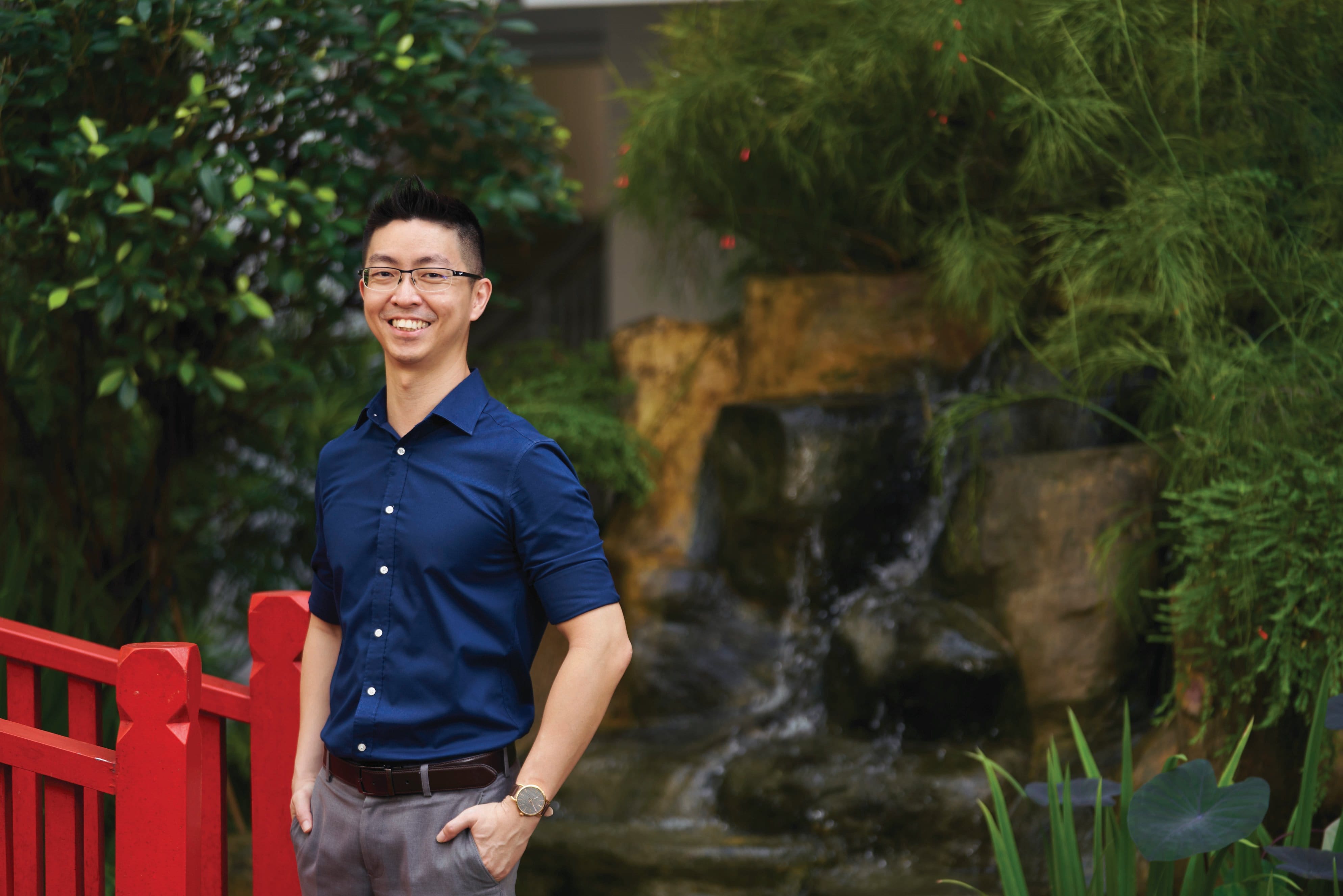 Smiling man in blue shirt and glasses stands by a red railing with a waterfall and green plants behind him.
