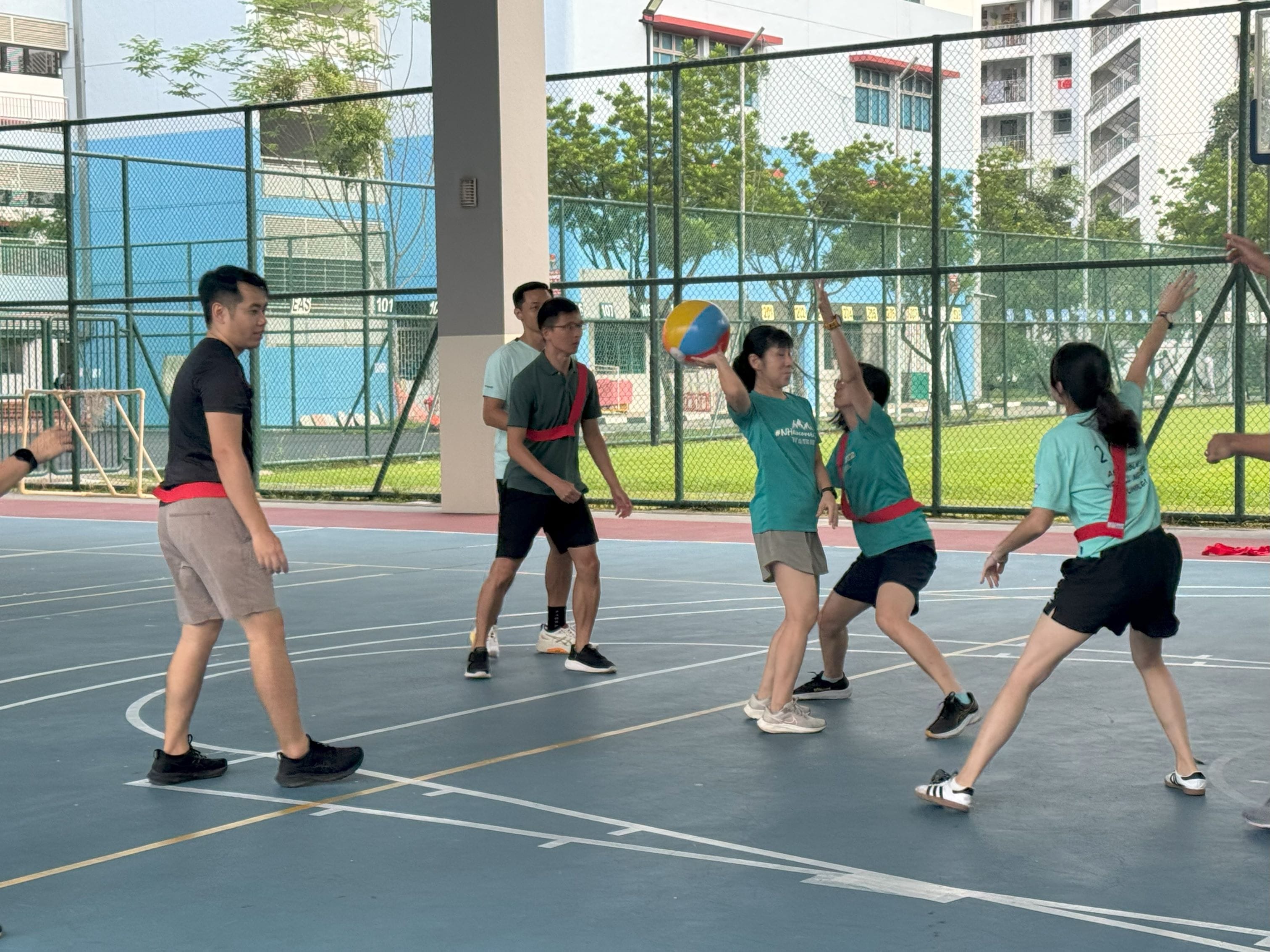 Several people play a ball game on an outdoor court, with one person holding a colorful ball overhead.