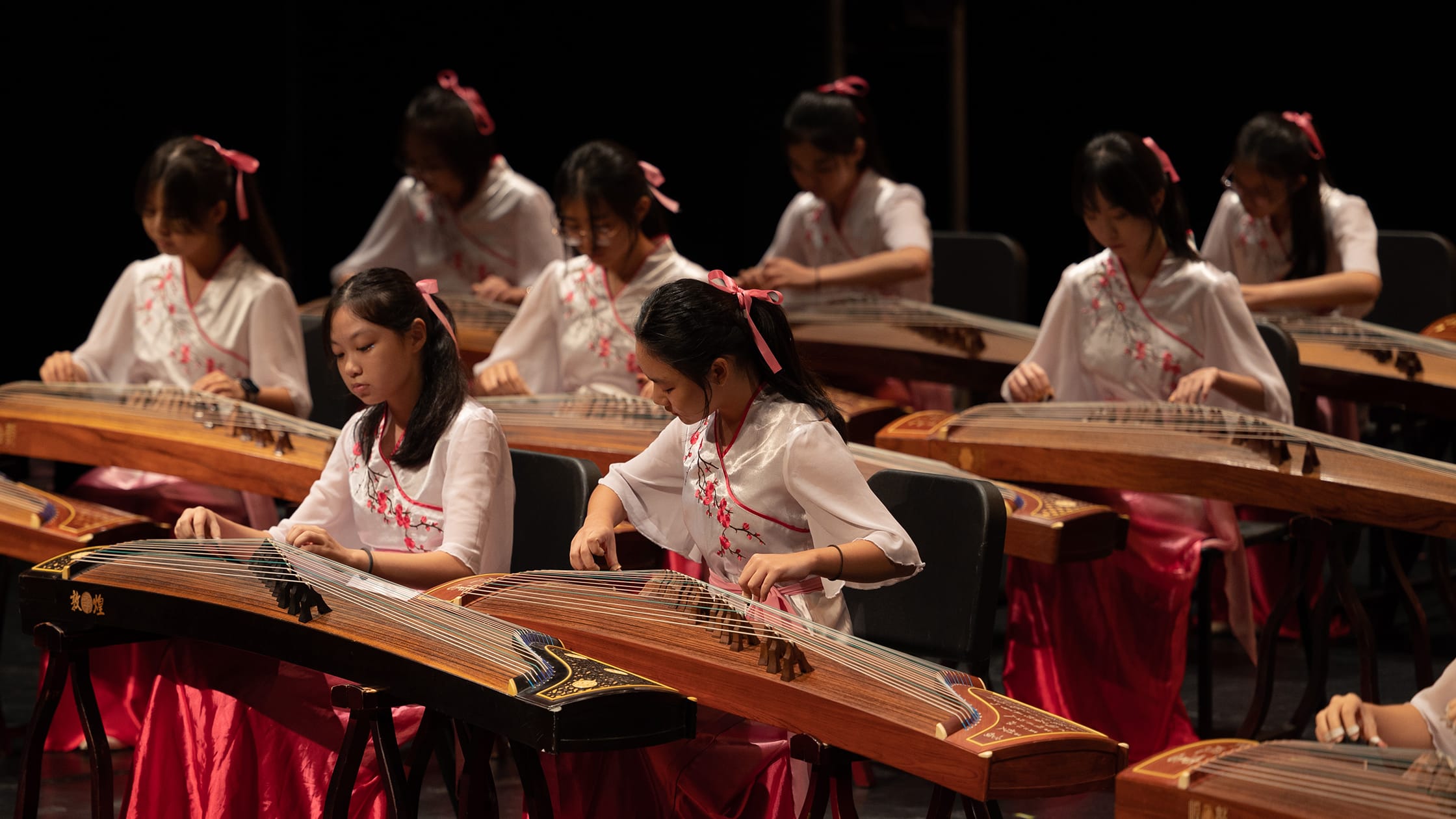 Young girls in white and pink outfits play guzheng instruments on a dark stage during a group performance.