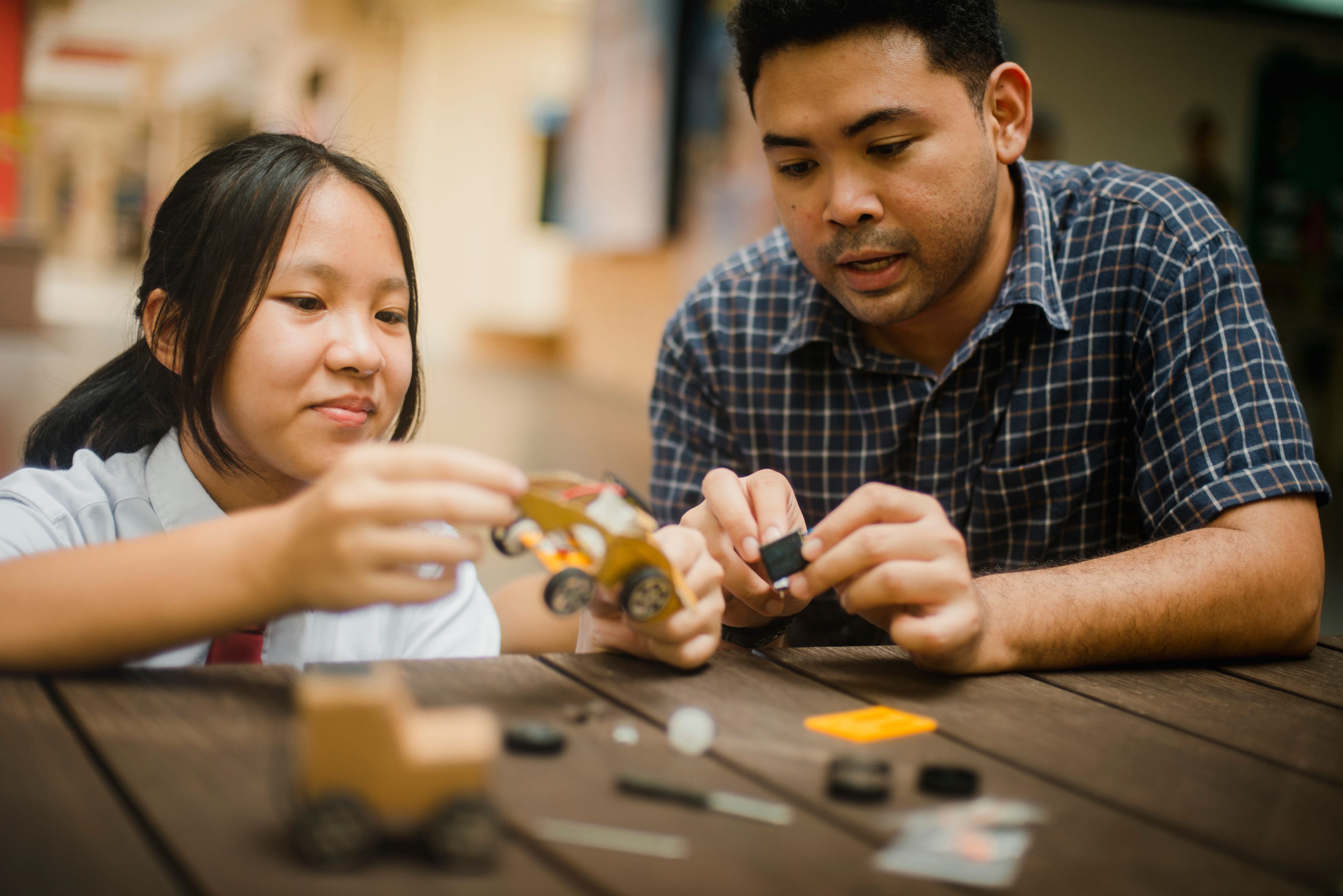 A man and a girl sit at a wooden table assembling a small yellow toy car together.