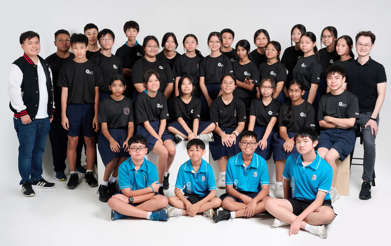 A large group of students and two adults pose for a formal photo, most wearing black shirts, four in blue polo shirts.