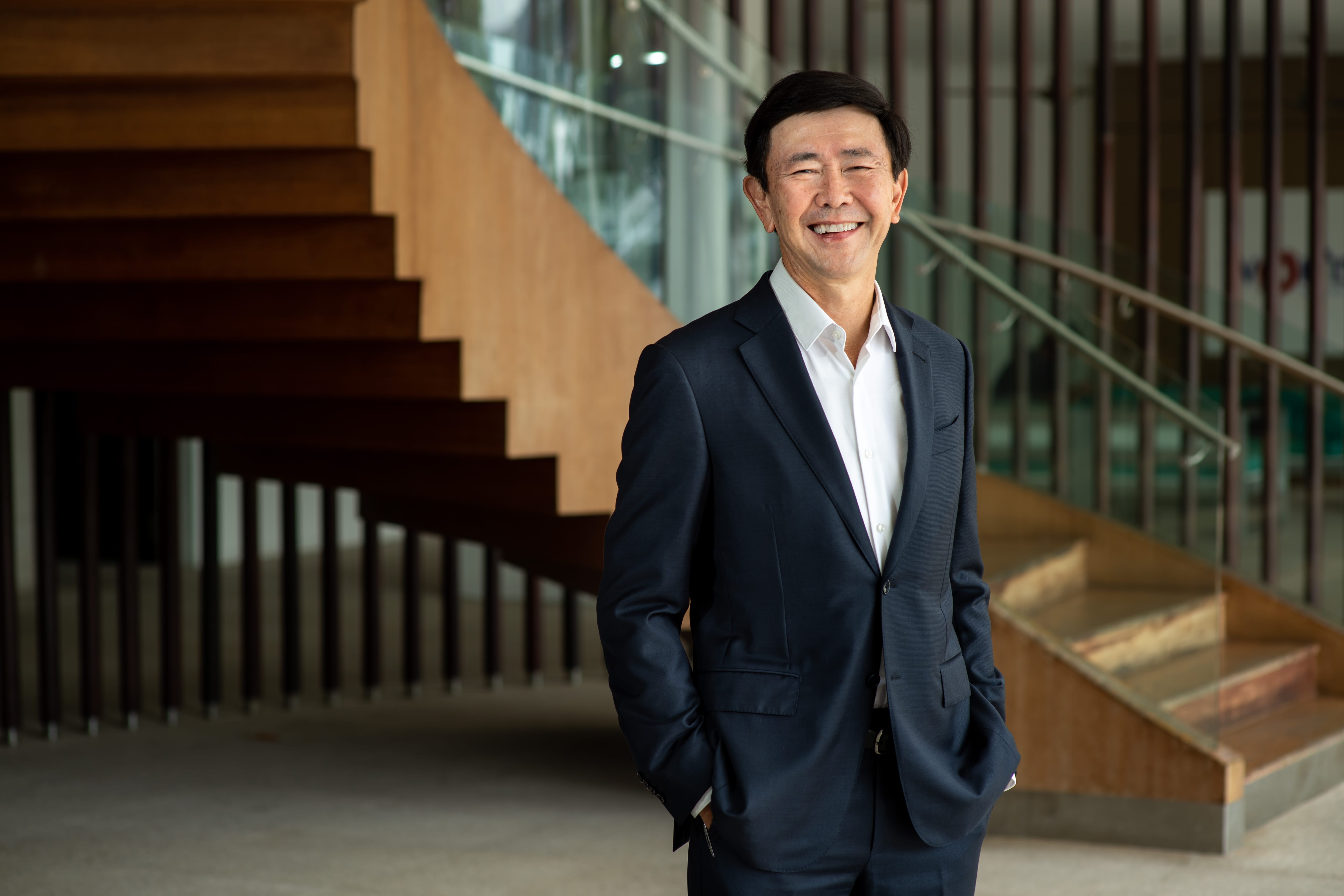 A smiling middle-aged Asian man in a navy suit and white shirt stands in front of wooden stairs indoors.