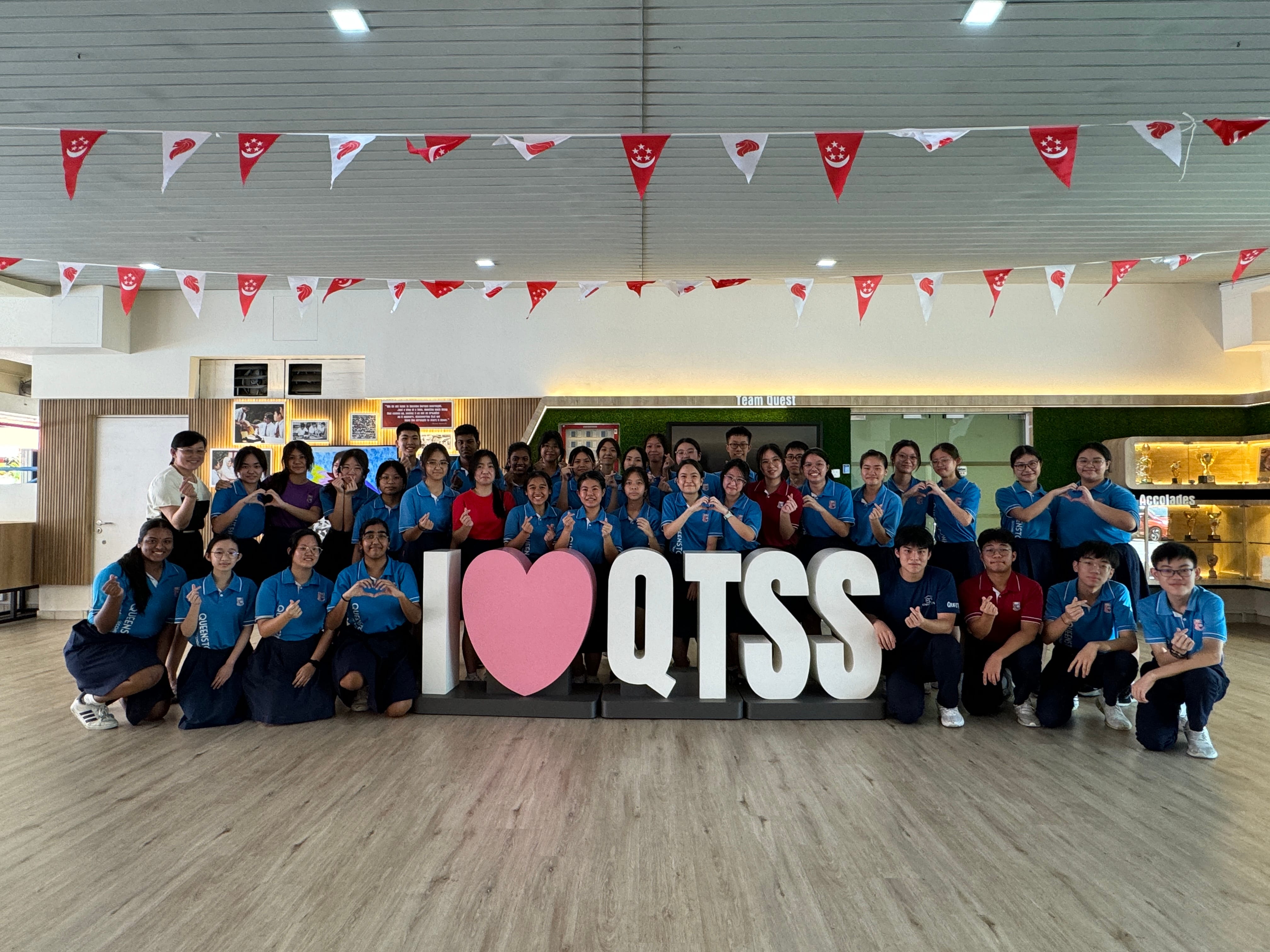 A large group of students in blue uniforms pose behind an "I Love QTSS" sign with Singapore flag bunting overhead.