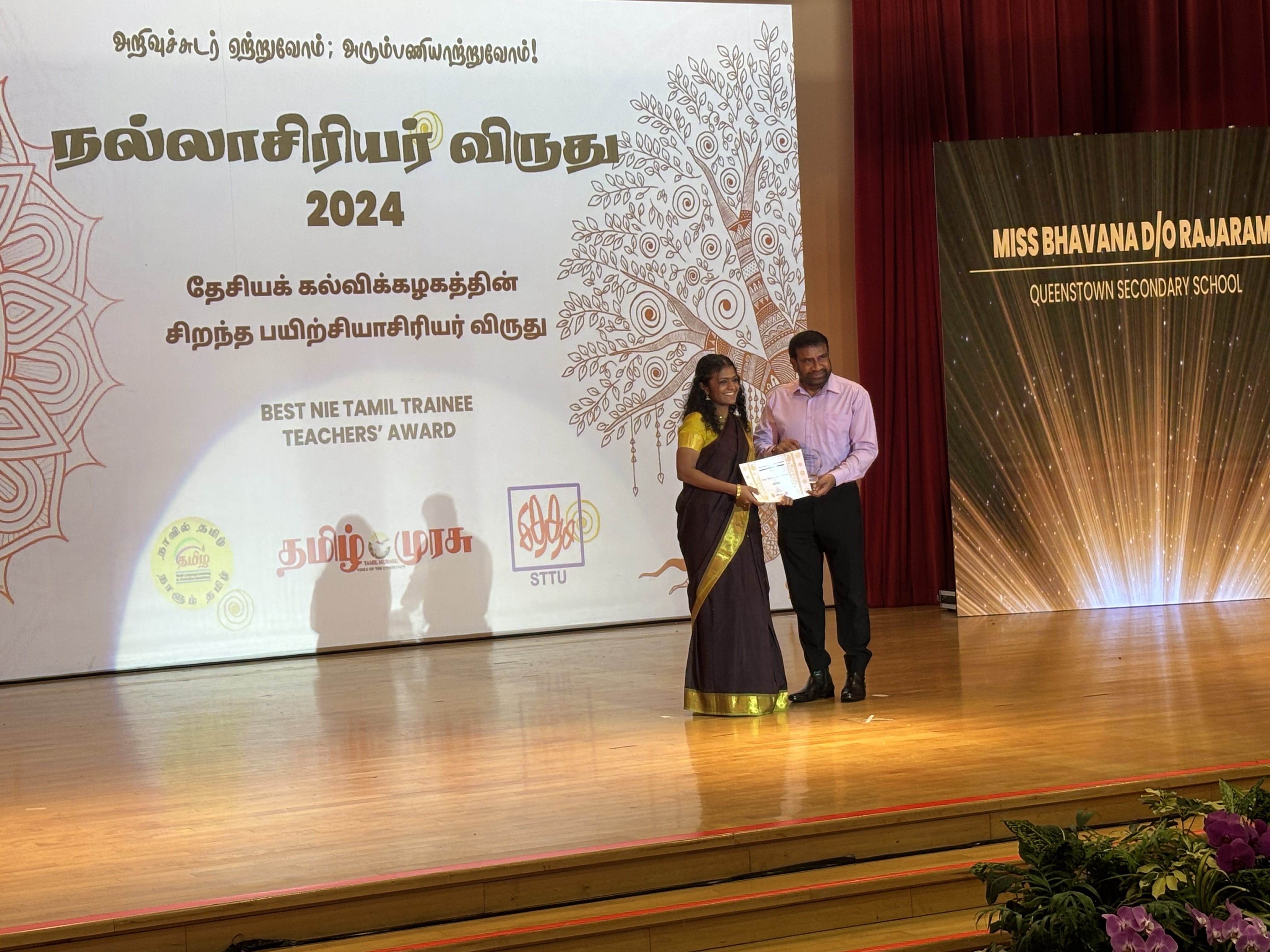 Two people on stage at the 2024 Best NIE Tamil Trainee Teachers Award ceremony, holding a certificate.