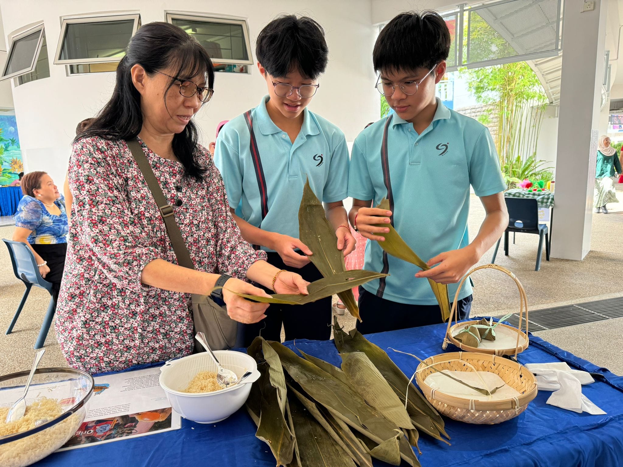 A woman and two students in blue uniforms wrap rice dumplings in bamboo leaves at a table.