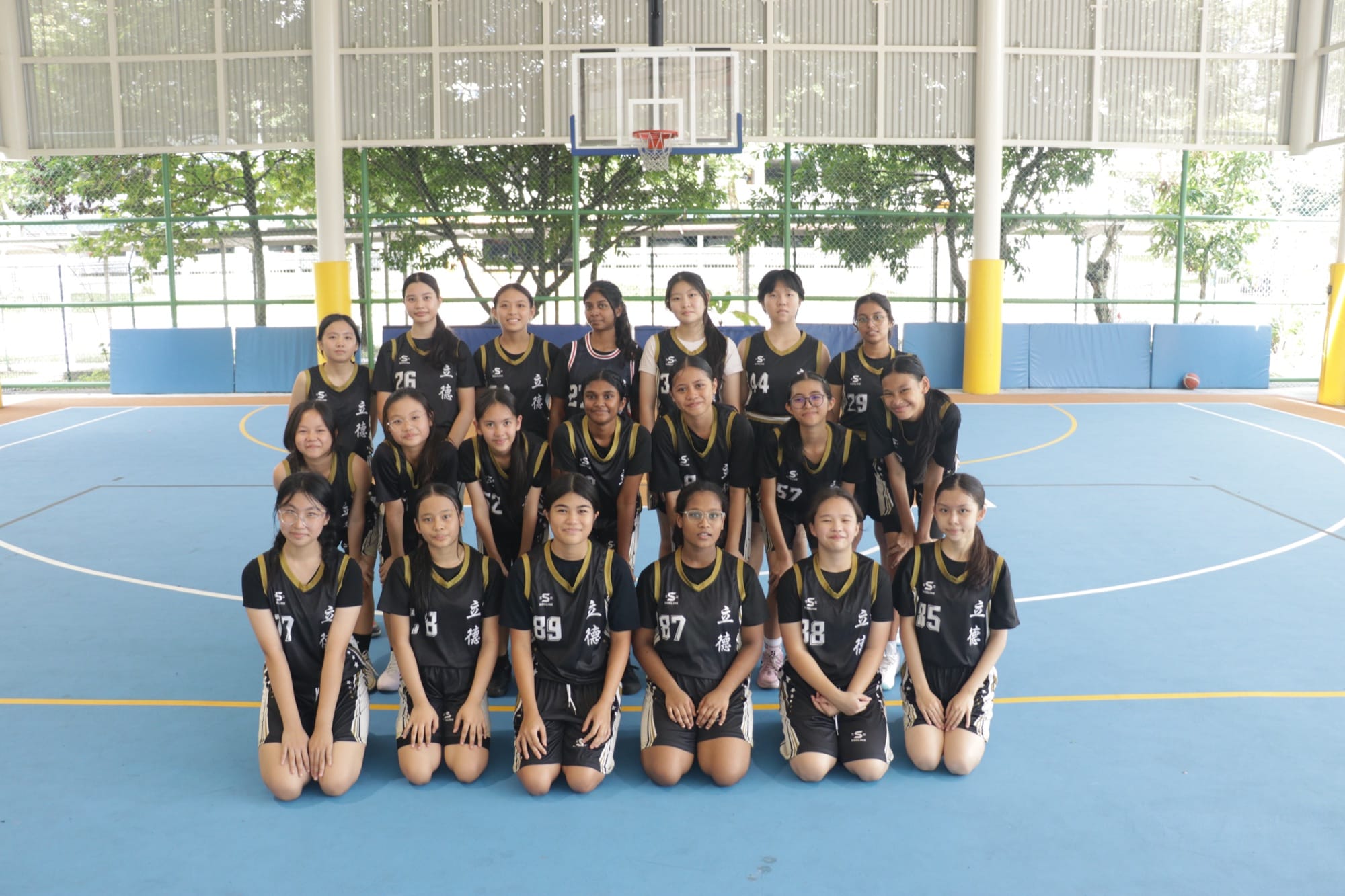 The Basketball (Girls) team in black and gold jerseys, on the school's outdoor basketball court in 2025.