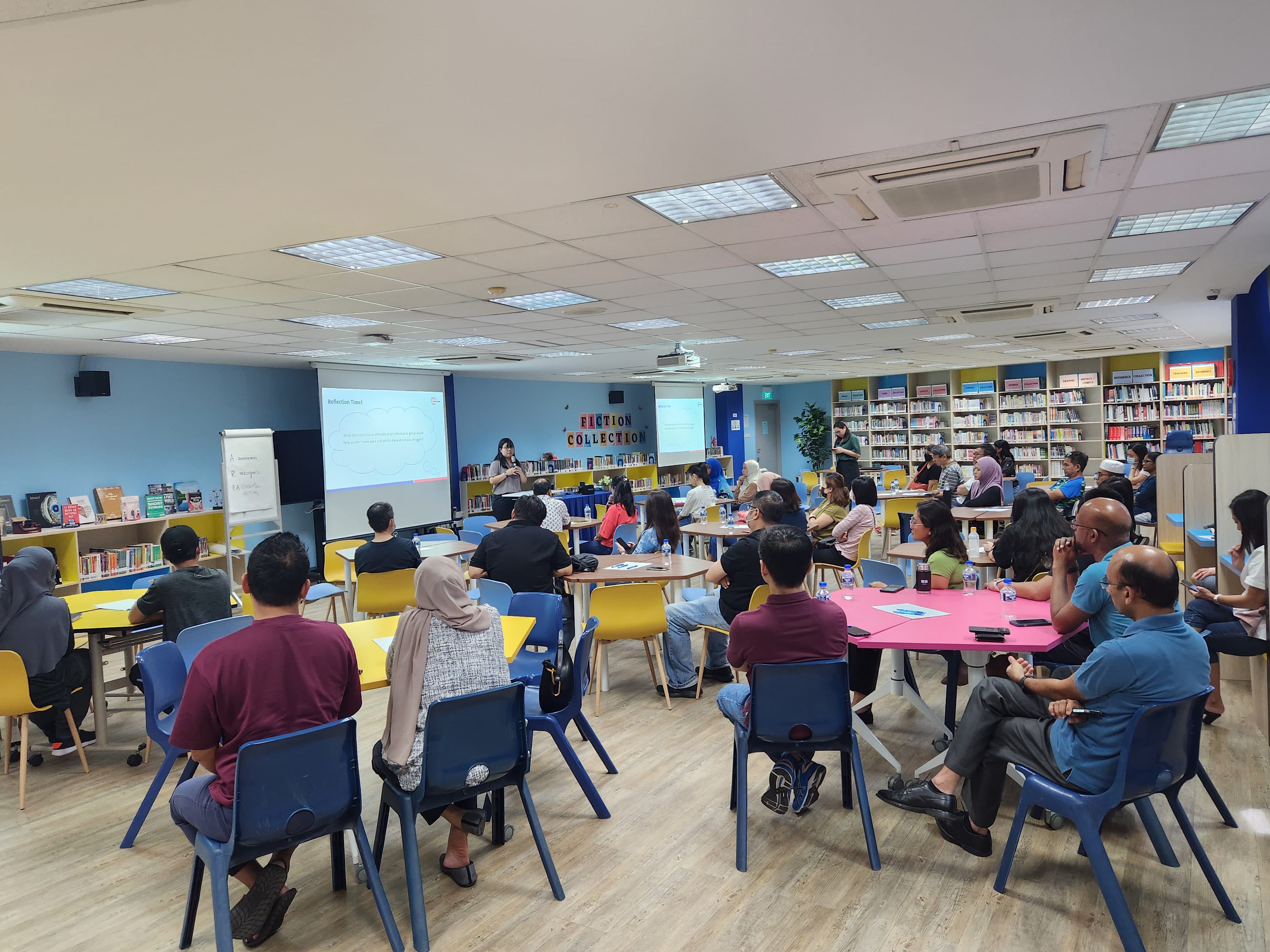 A presenter stands before a large group seated at colourful tables in a library with bookshelves and a projected screen.