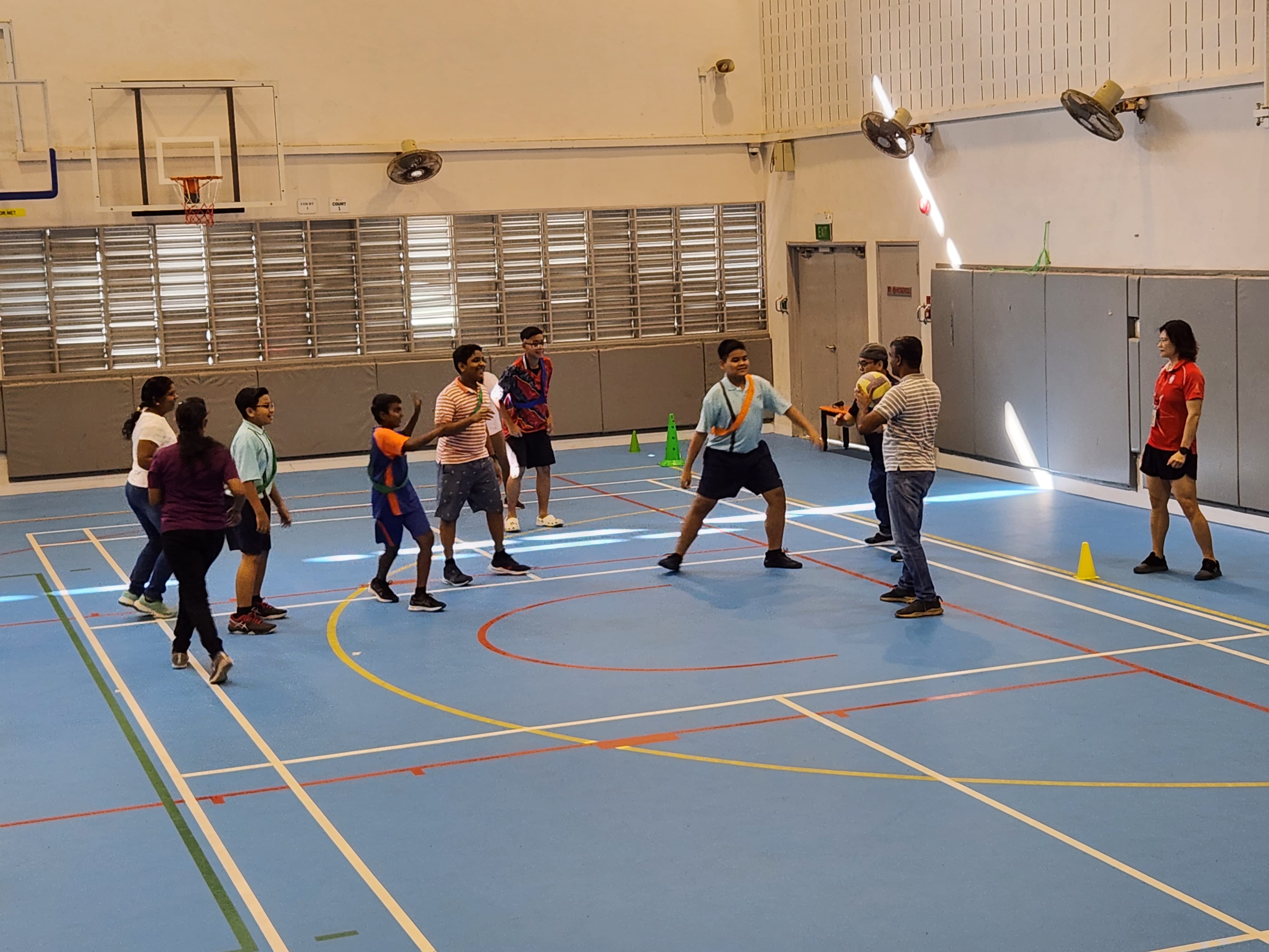 A group of children and adults playing a ball game on a blue indoor sports court with cones and a basketball hoop.