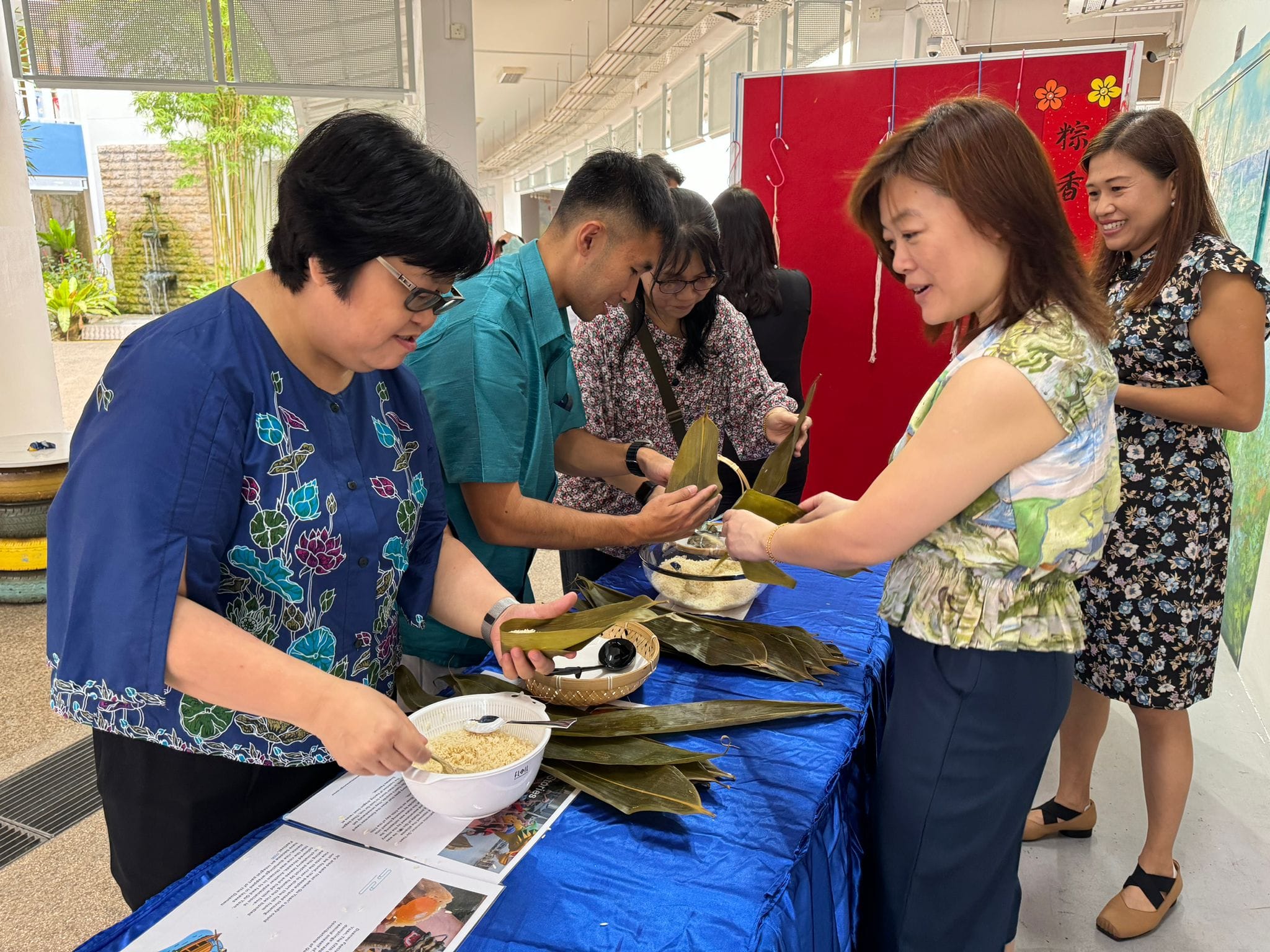 Four people wrap zongzi rice dumplings using bamboo leaves and rice at a blue-covered table indoors.