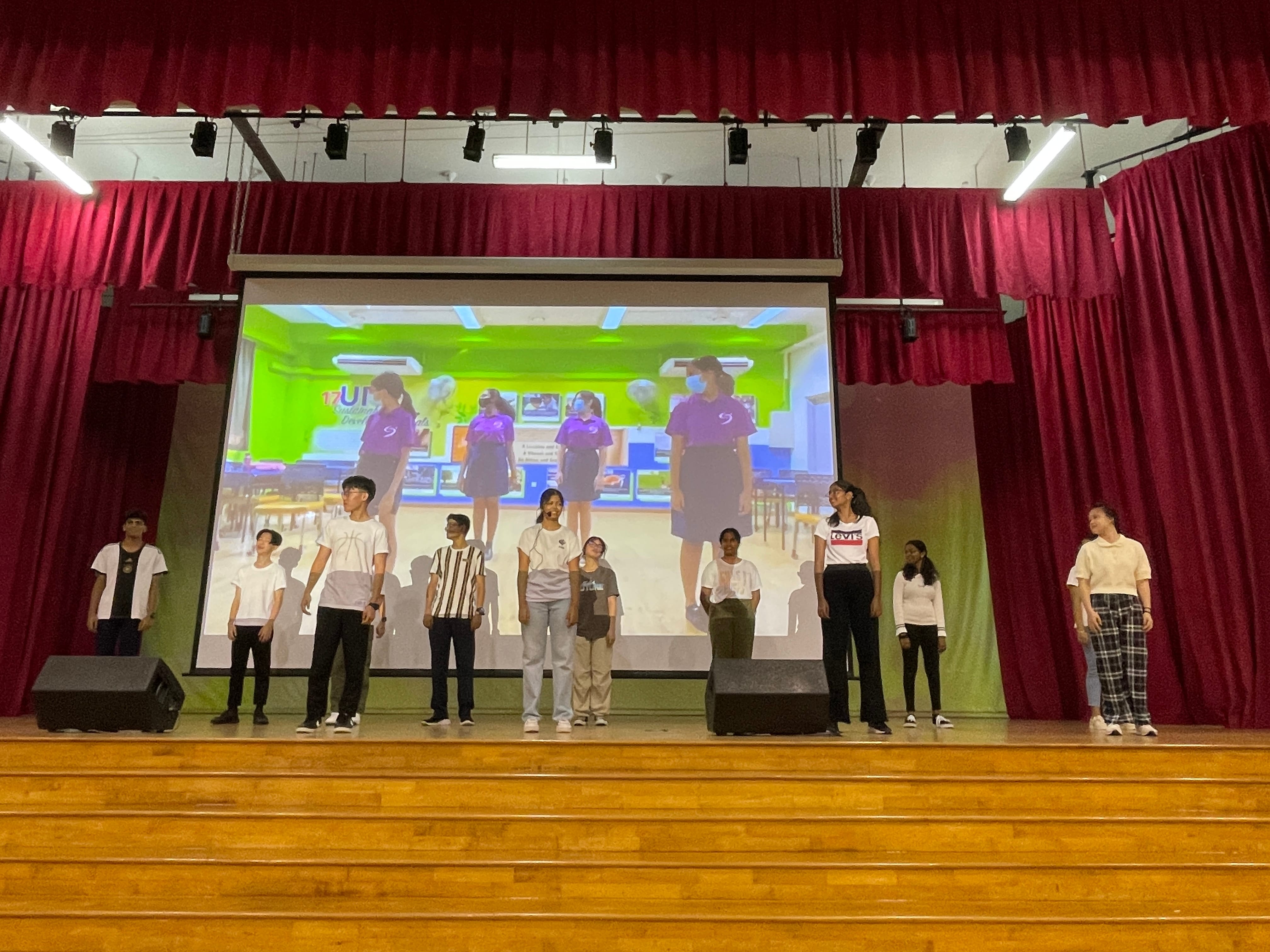 Students stand on a wooden stage in front of red curtains and a projected screen showing purple-uniformed students.