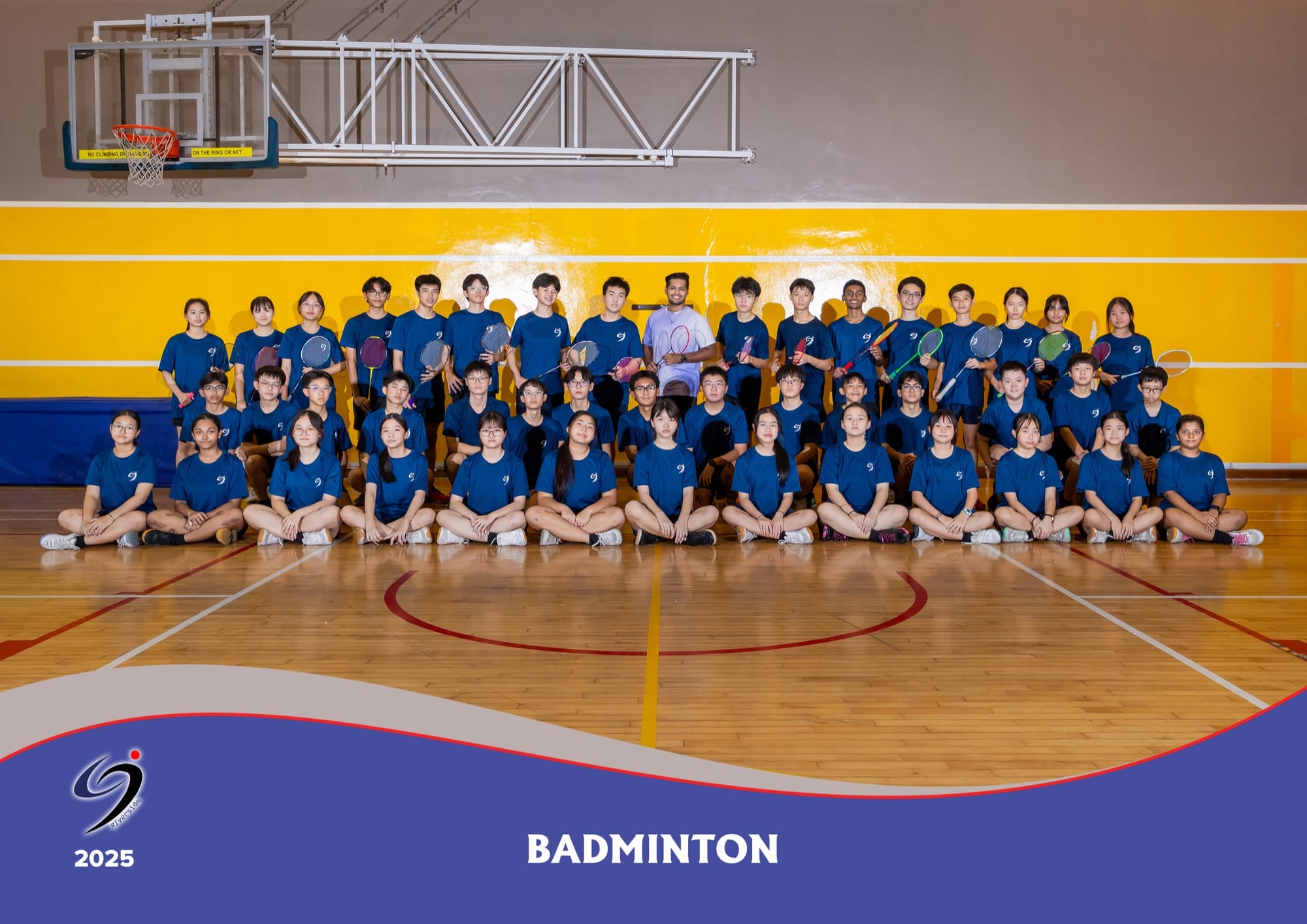 Badminton team members in blue sports attire holding racquets, with their teacher-in-charge, in the indoor sports hall in 2025.
