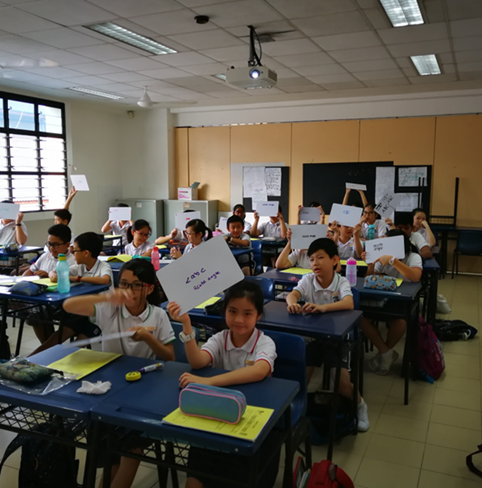 Students in school uniforms sit at blue desks holding up white papers with writing in a classroom.