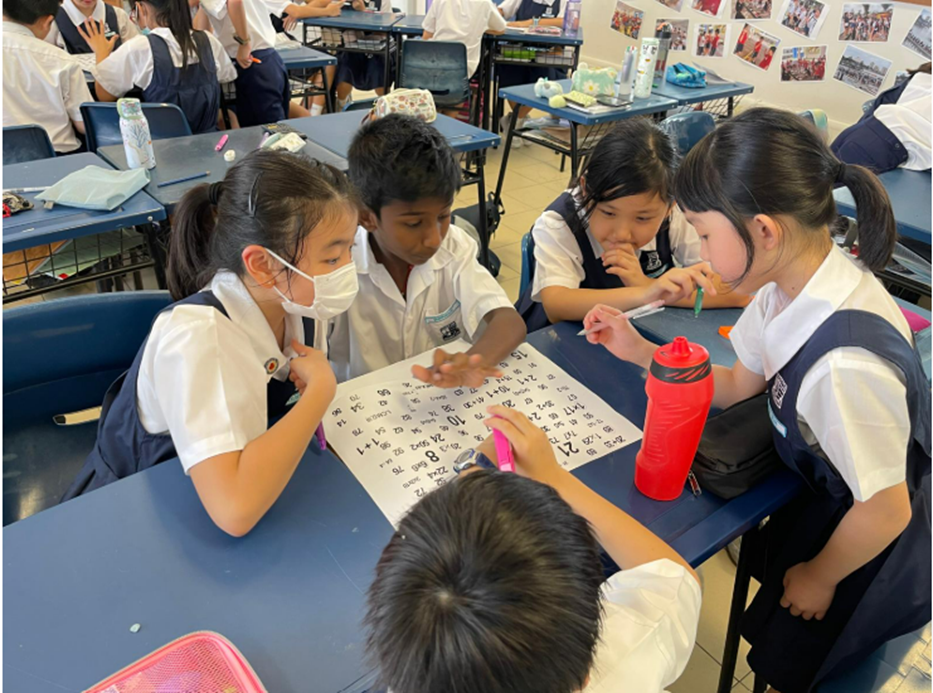 Five uniformed primary school students sit around a desk working together on a math numbers sheet in a classroom.