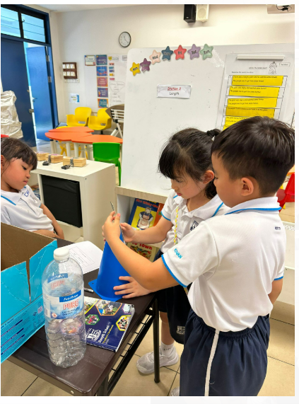 Two young students in white uniforms use a blue funnel at a classroom table with a water bottle nearby.