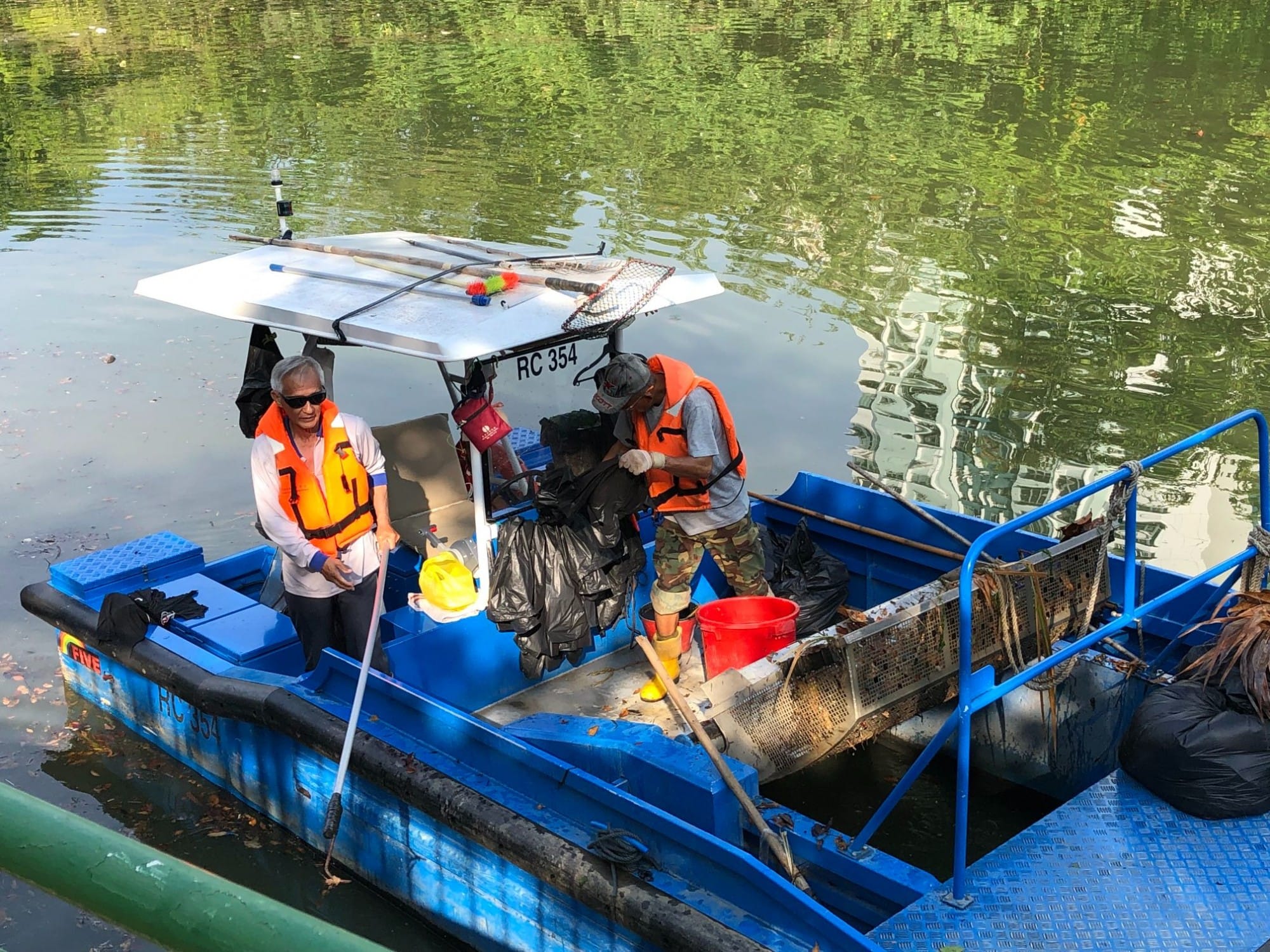 Two workers in orange life vests collect trash on a blue boat labeled RC 354 on a green river.