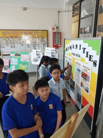 Students in blue uniforms looking at a colorful Humanities Corner display board in a school hallway.