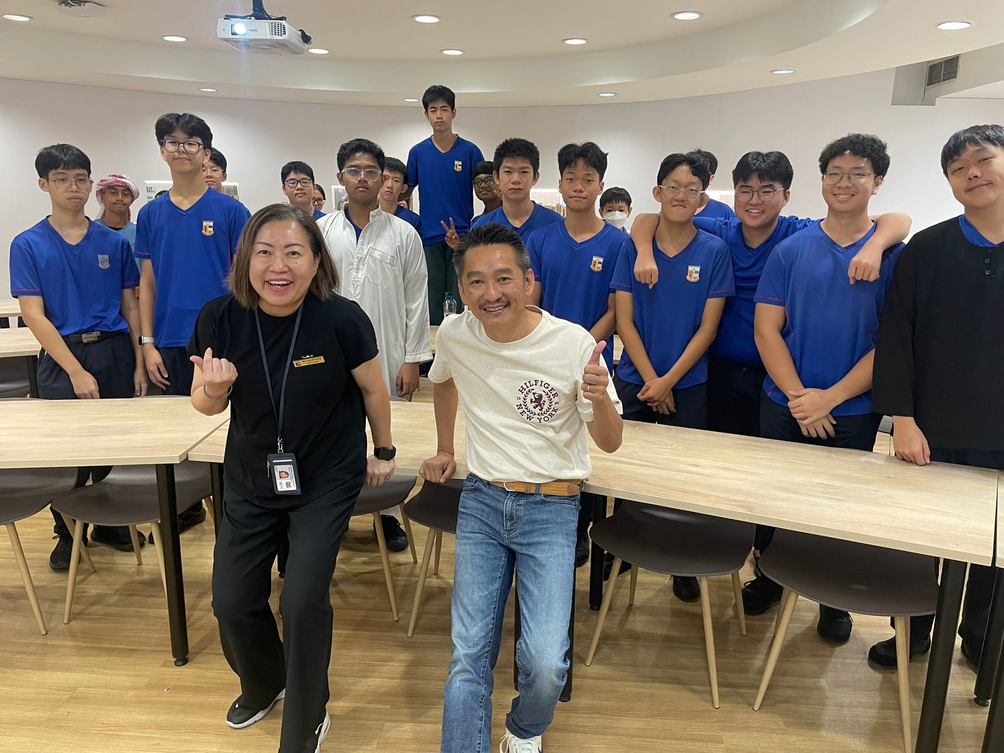 Two adults pose with thumbs up in front of a large group of teenage boys in blue school uniforms indoors.