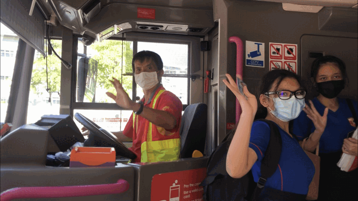A masked bus driver in a yellow vest waves alongside two masked passengers waving inside a bus.
