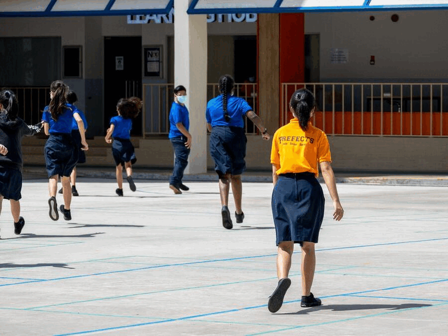 Students in blue uniforms run across a school courtyard, watched by a prefect in an orange shirt.