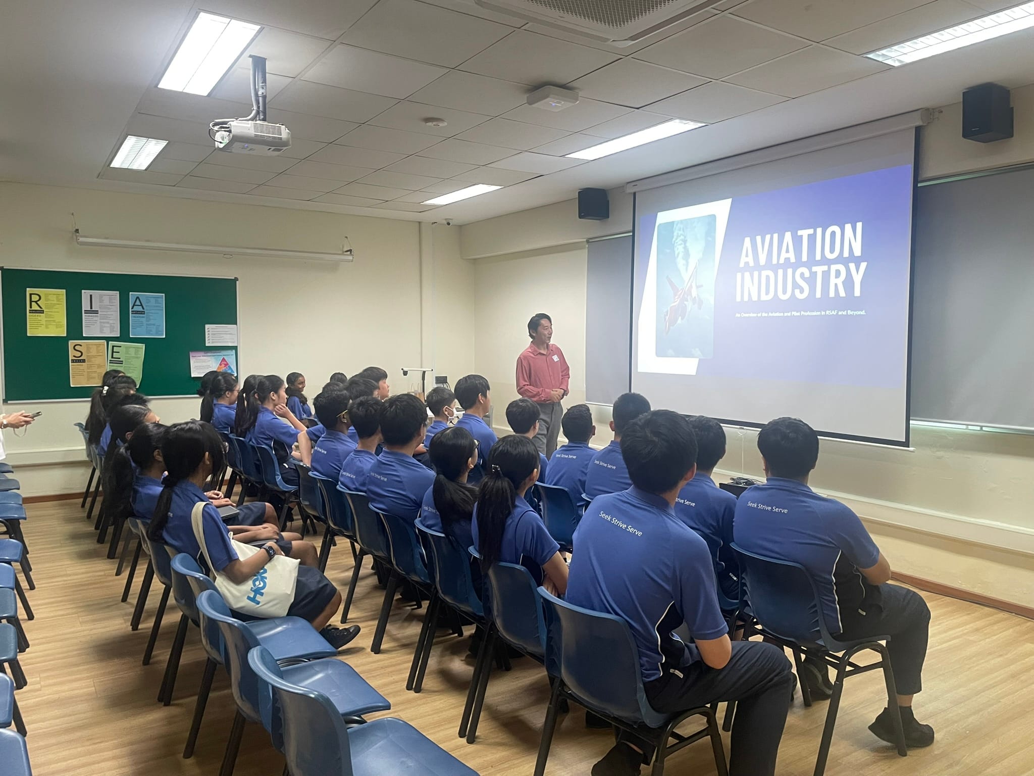 Students in blue uniforms sit in a classroom watching a presenter beside a screen showing "Aviation Industry."