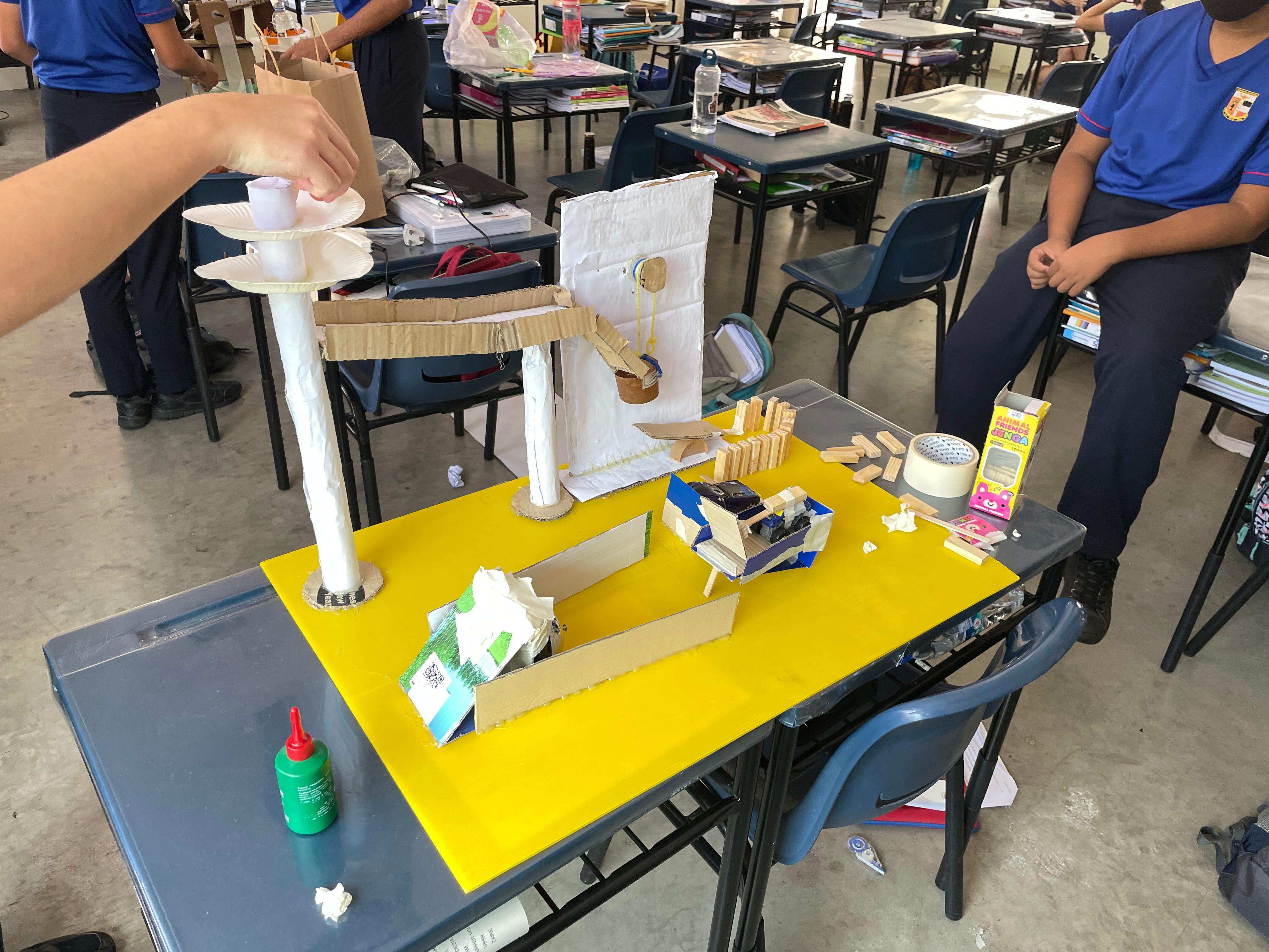 Students in a classroom build a cardboard marble run on a yellow board, with glue and blocks nearby.