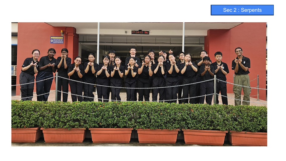 A group of students in black uniforms pose together outdoors in front of a red building, making hand gestures.