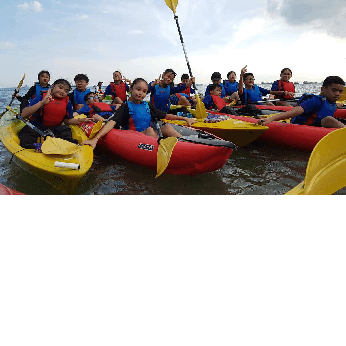 A group of children wearing red life jackets sit in red and yellow kayaks on calm water, posing with paddles.