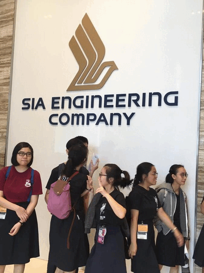 Several young visitors with lanyards stand in front of a SIA Engineering Company sign with a gold logo.