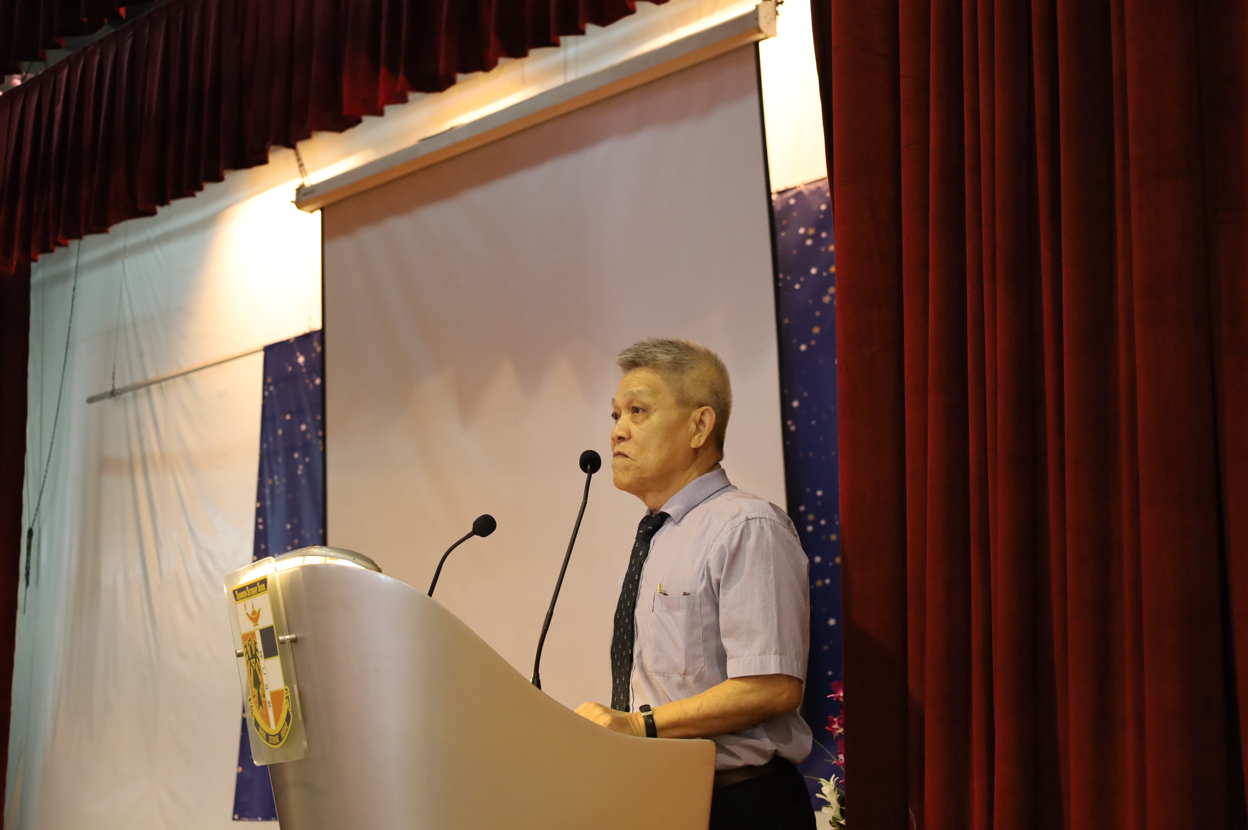 An older man in a light blue shirt and tie speaks at a podium with two microphones on a stage.