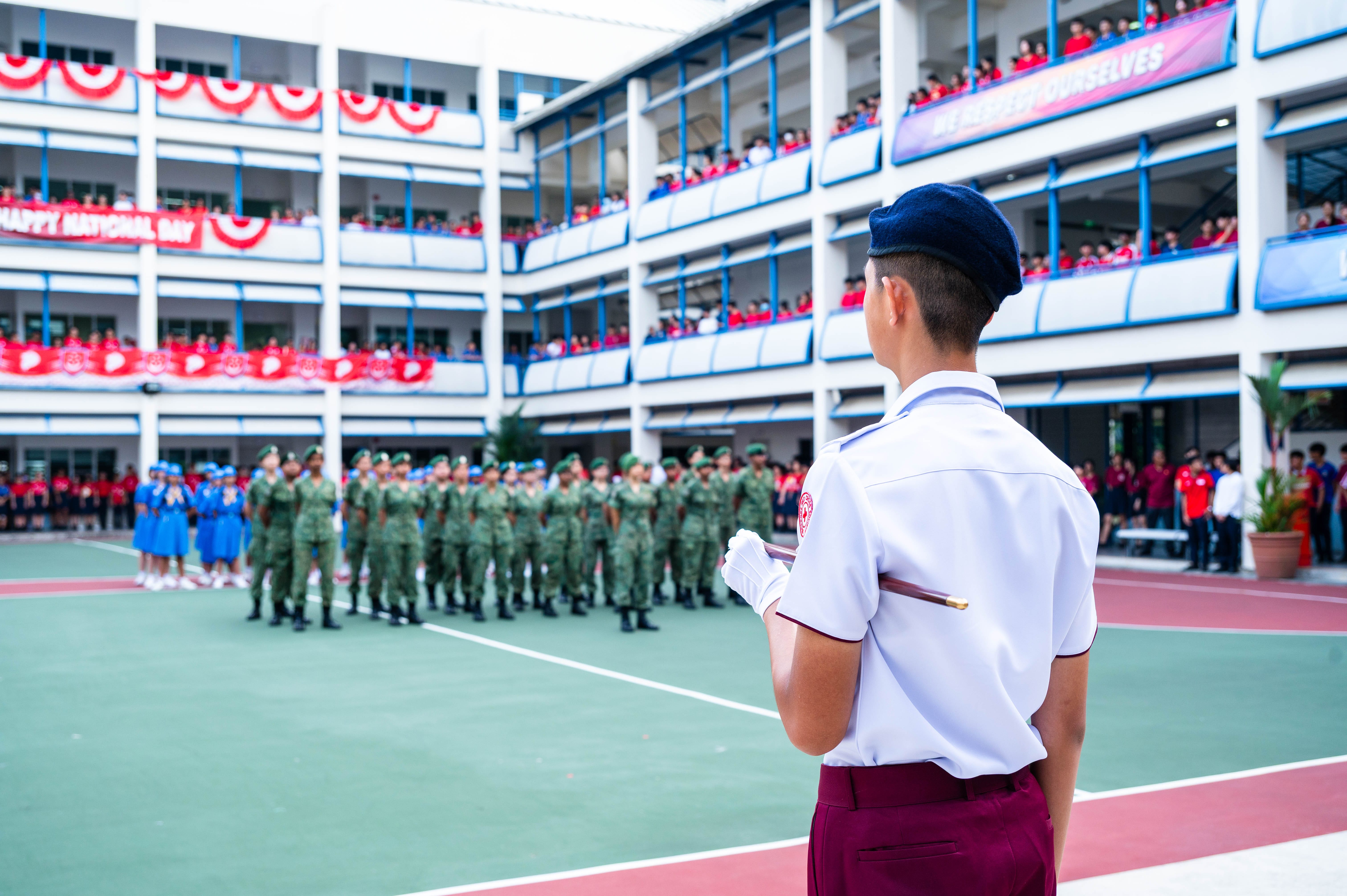 A uniformed cadet in a blue beret faces rows of soldiers on a school courtyard during a Happy National Day ceremony.