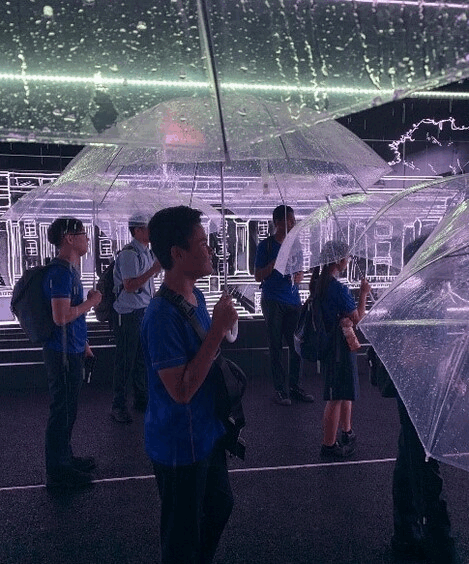 People holding clear umbrellas stand inside a dark immersive installation with glowing neon building outlines.