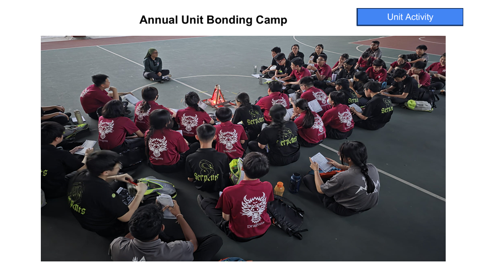 Students in matching red and black shirts sit in a circle on a court during an Annual Unit Bonding Camp activity.