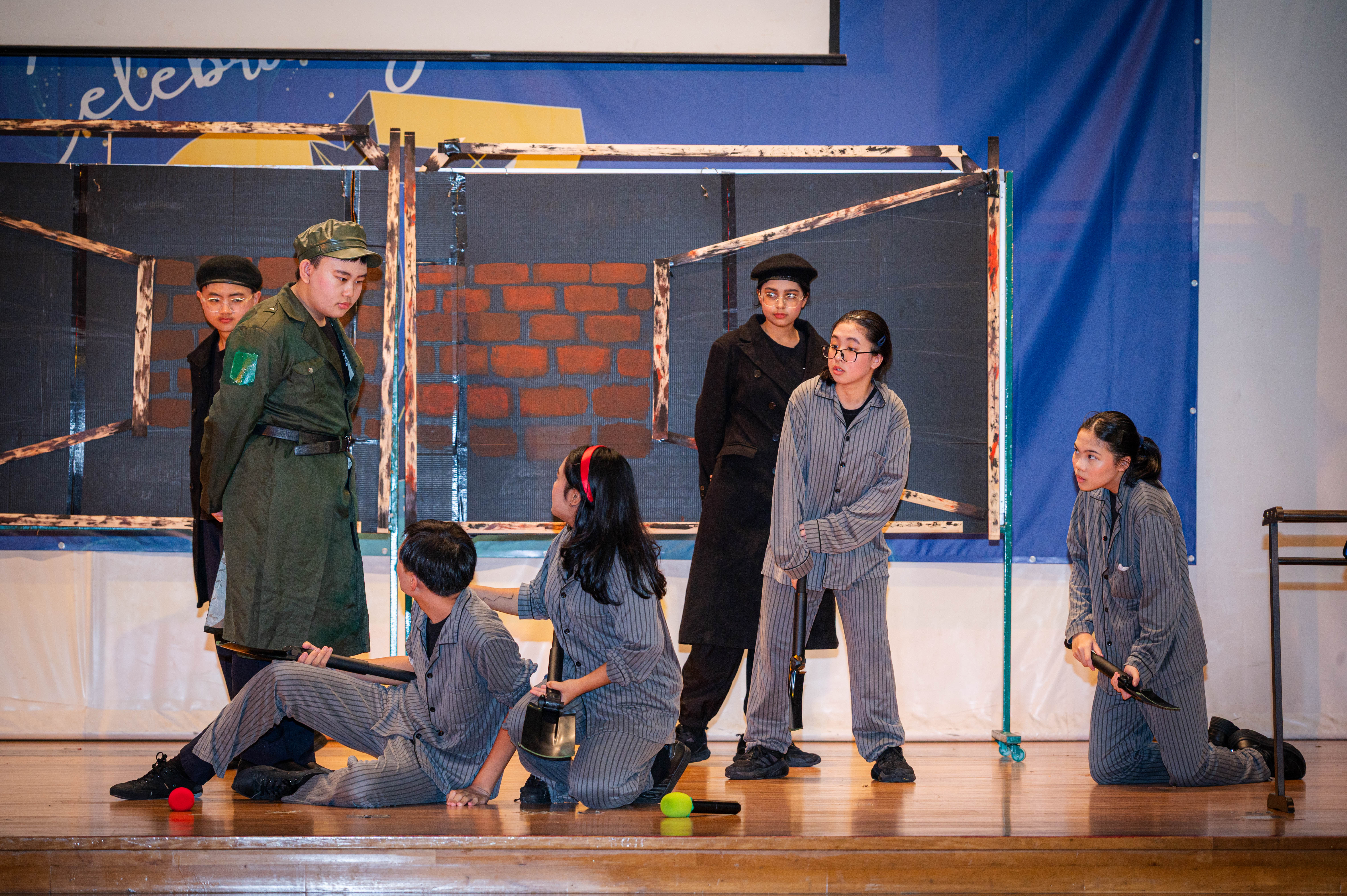 Students perform a stage play, some in striped prisoner costumes and one in a green military uniform, before a brick wall backdrop.