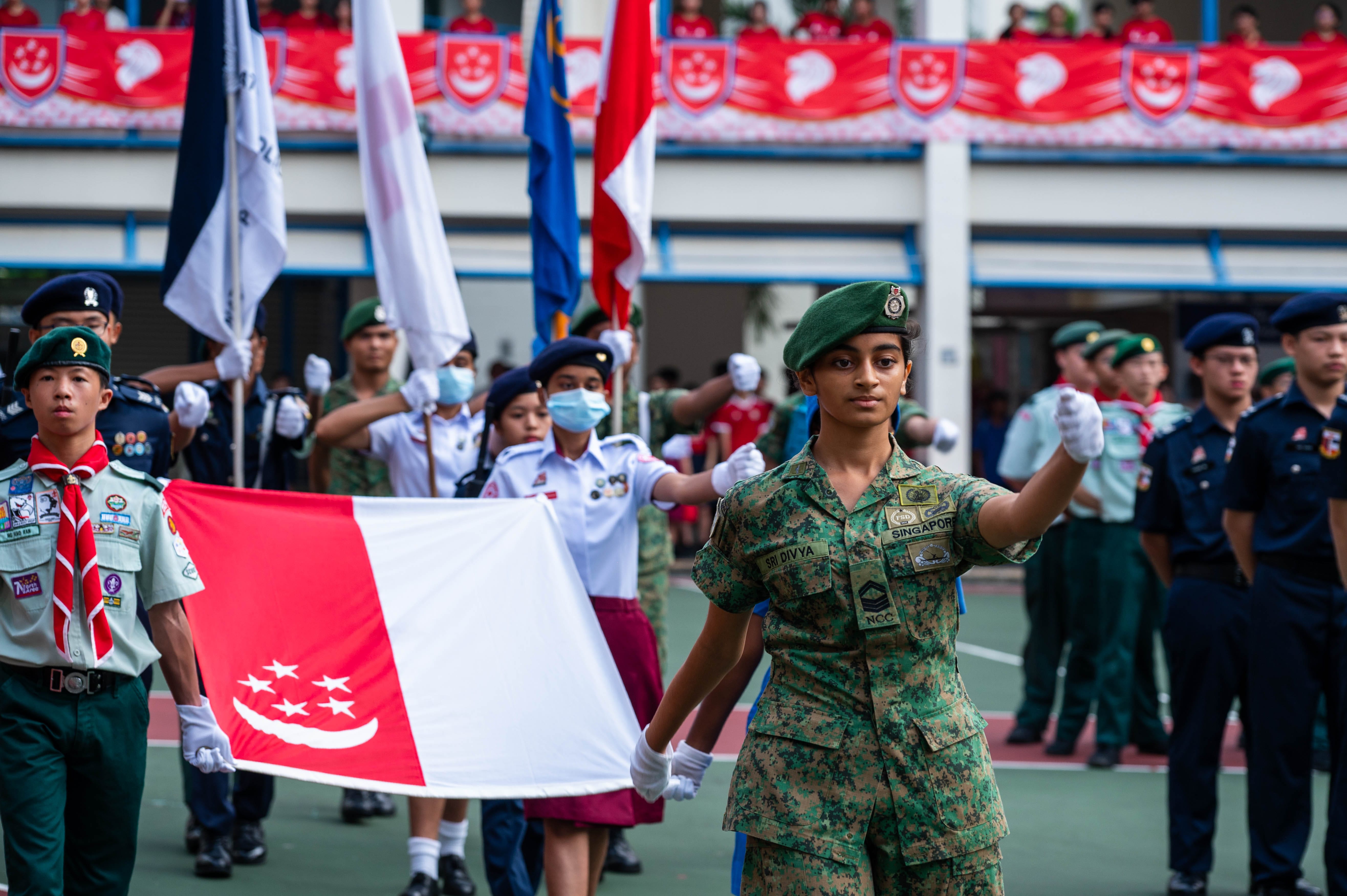 A girl in Singapore NCC uniform holds the Singapore flag during a parade with scouts and cadets behind her.