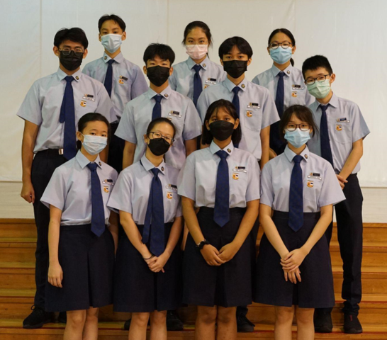 Twelve students in light blue uniforms and navy ties wearing face masks pose together in two rows.