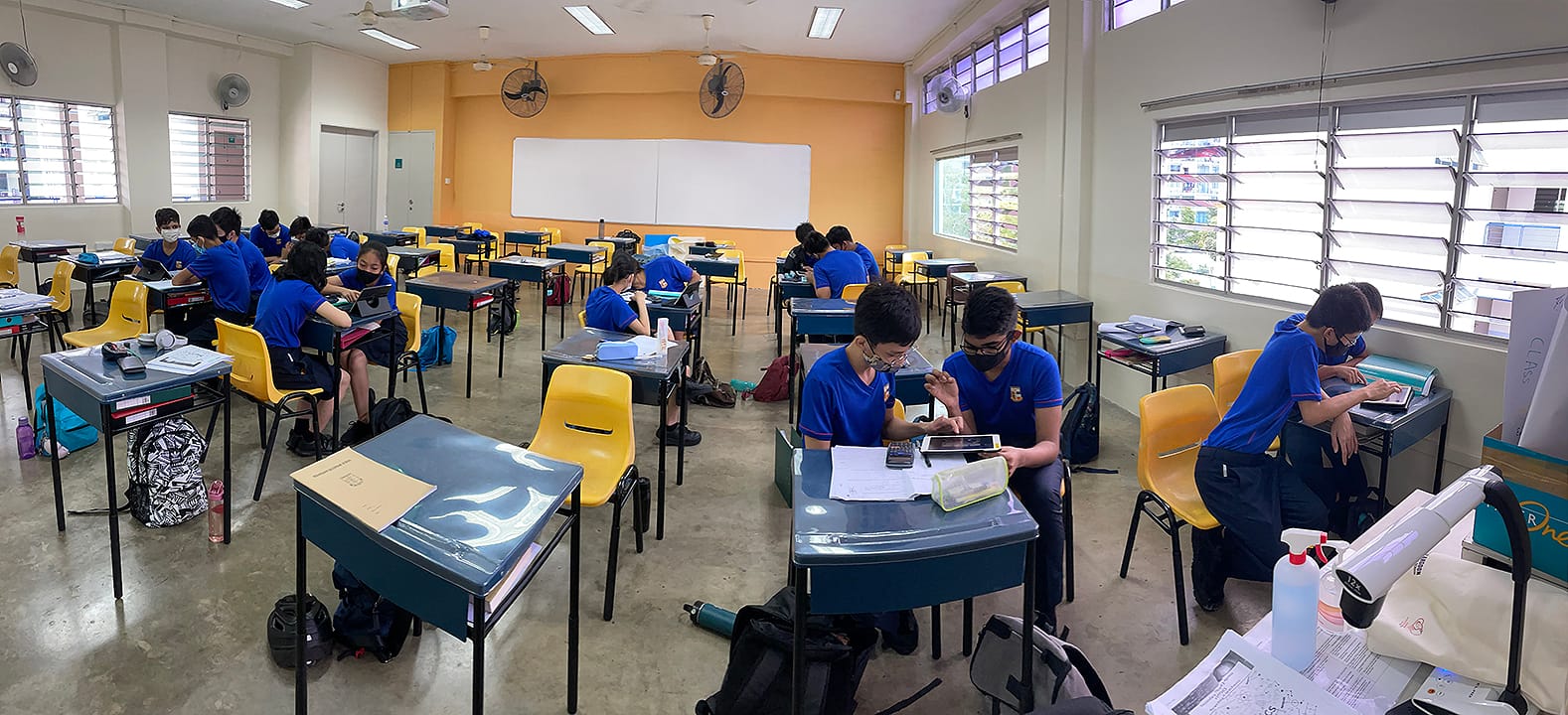 Students in blue uniforms and masks sit at spaced desks in a classroom with yellow chairs and a whiteboard.