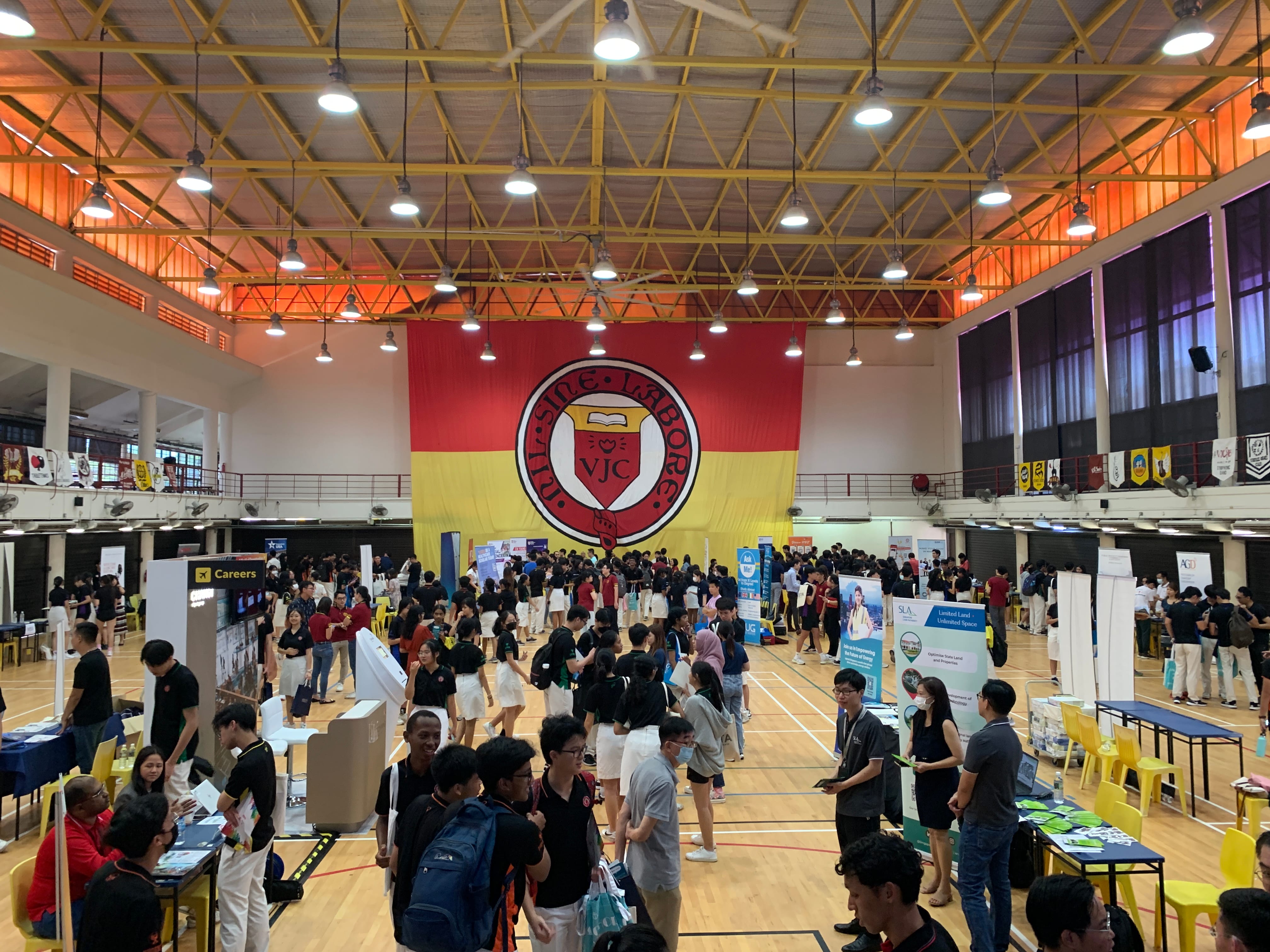 A crowded indoor careers fair in a gymnasium with a large red and yellow VJC school crest banner on the wall.