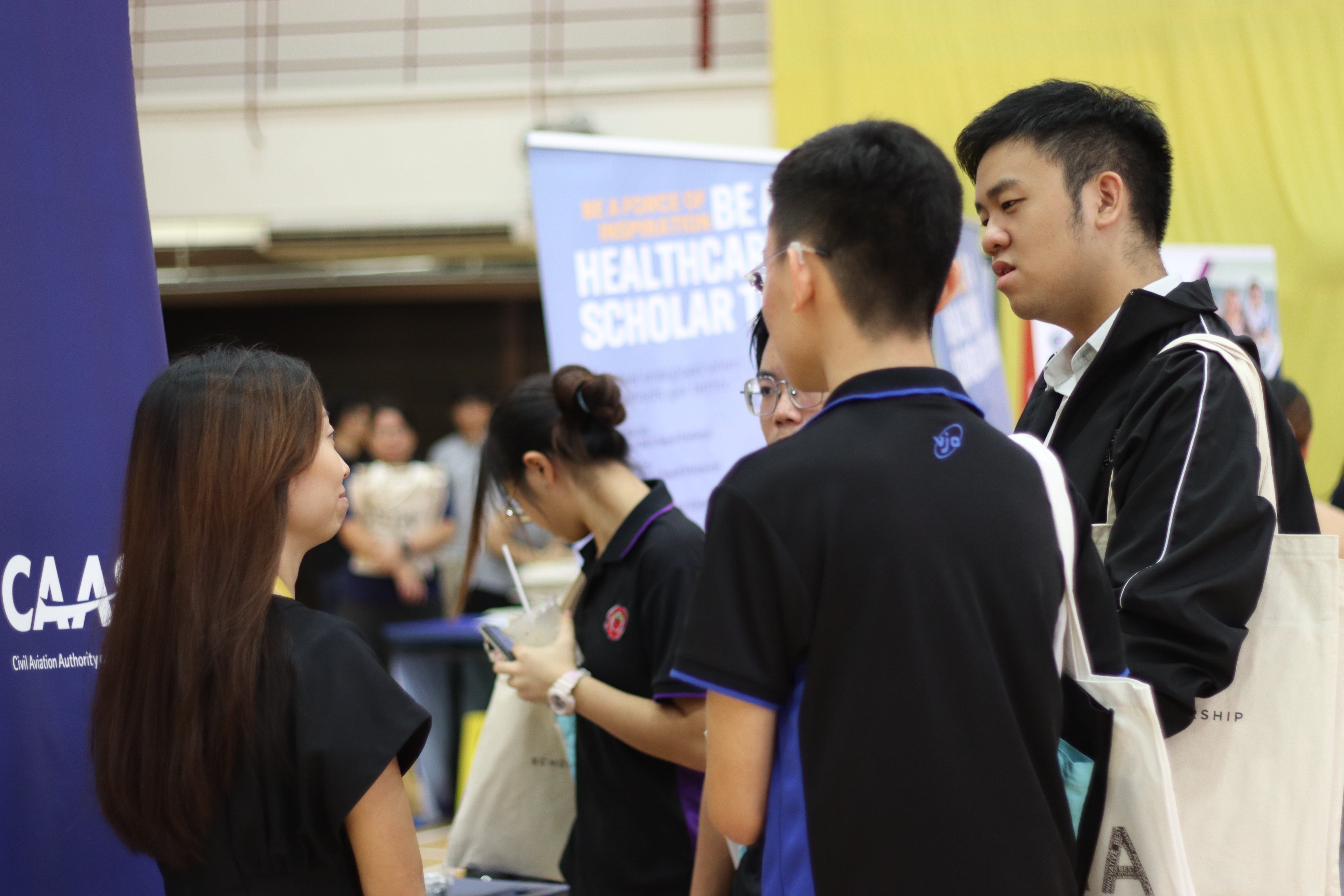 Four young people converse at a scholarship fair with a Healthcare Scholar banner and CAAS booth visible.