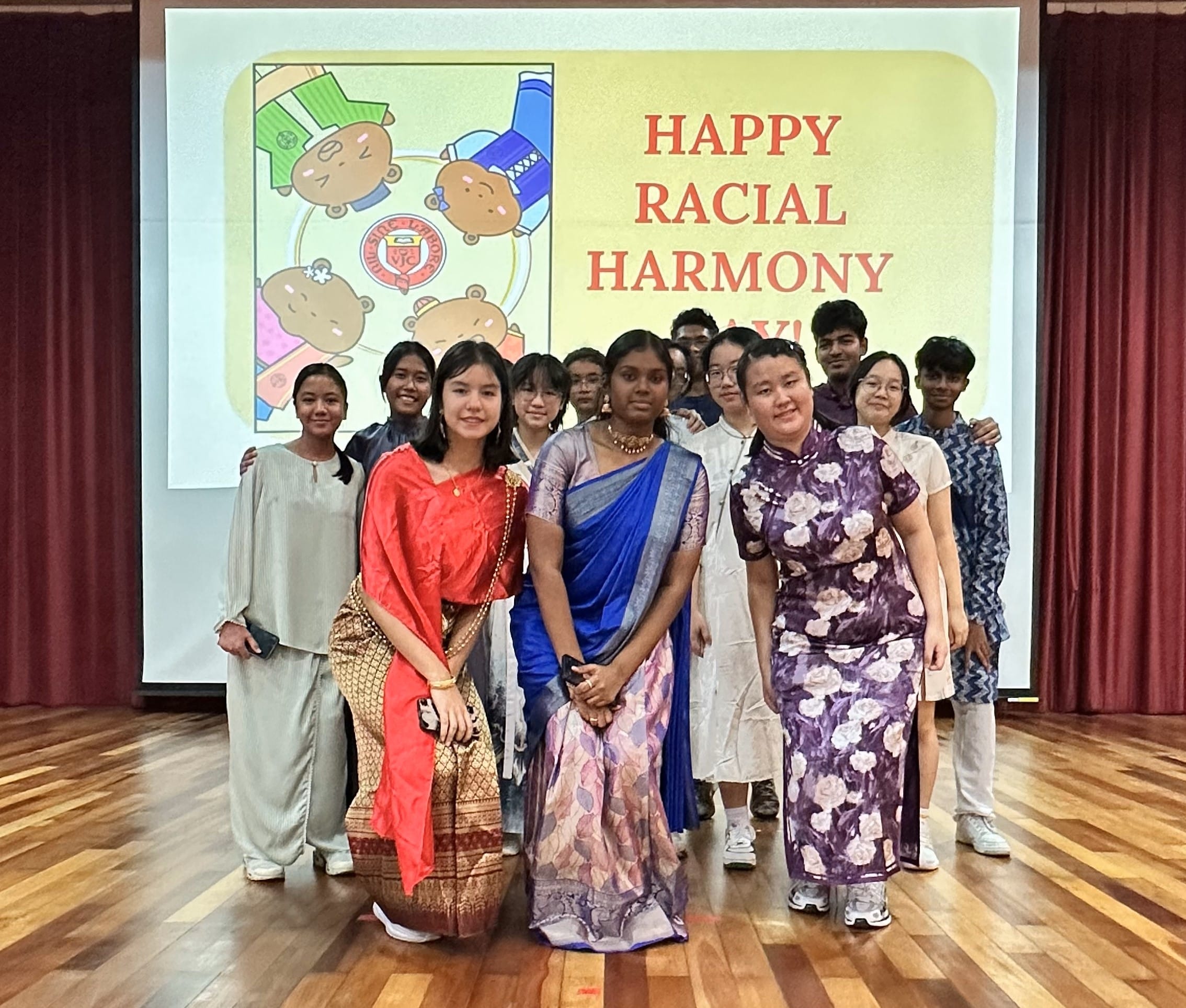 A group of students in traditional cultural outfits pose before a Happy Racial Harmony Day banner in a hall.