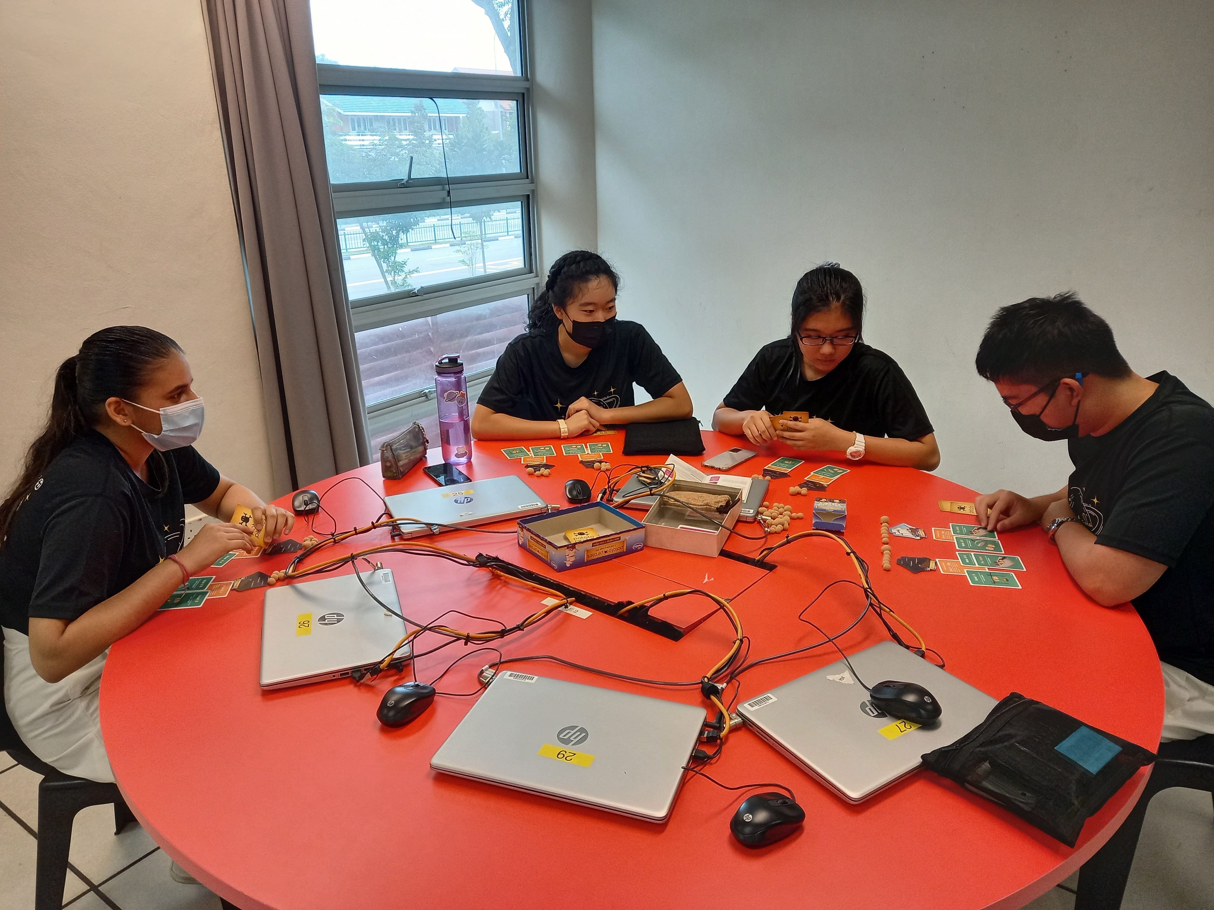 Four masked students sit around a red round table with HP laptops, cards, and small wooden beads.