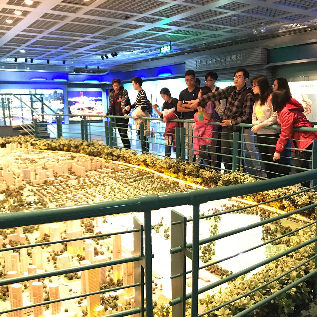People stand on a raised walkway viewing a large detailed scale model of a city with tiny buildings and trees.