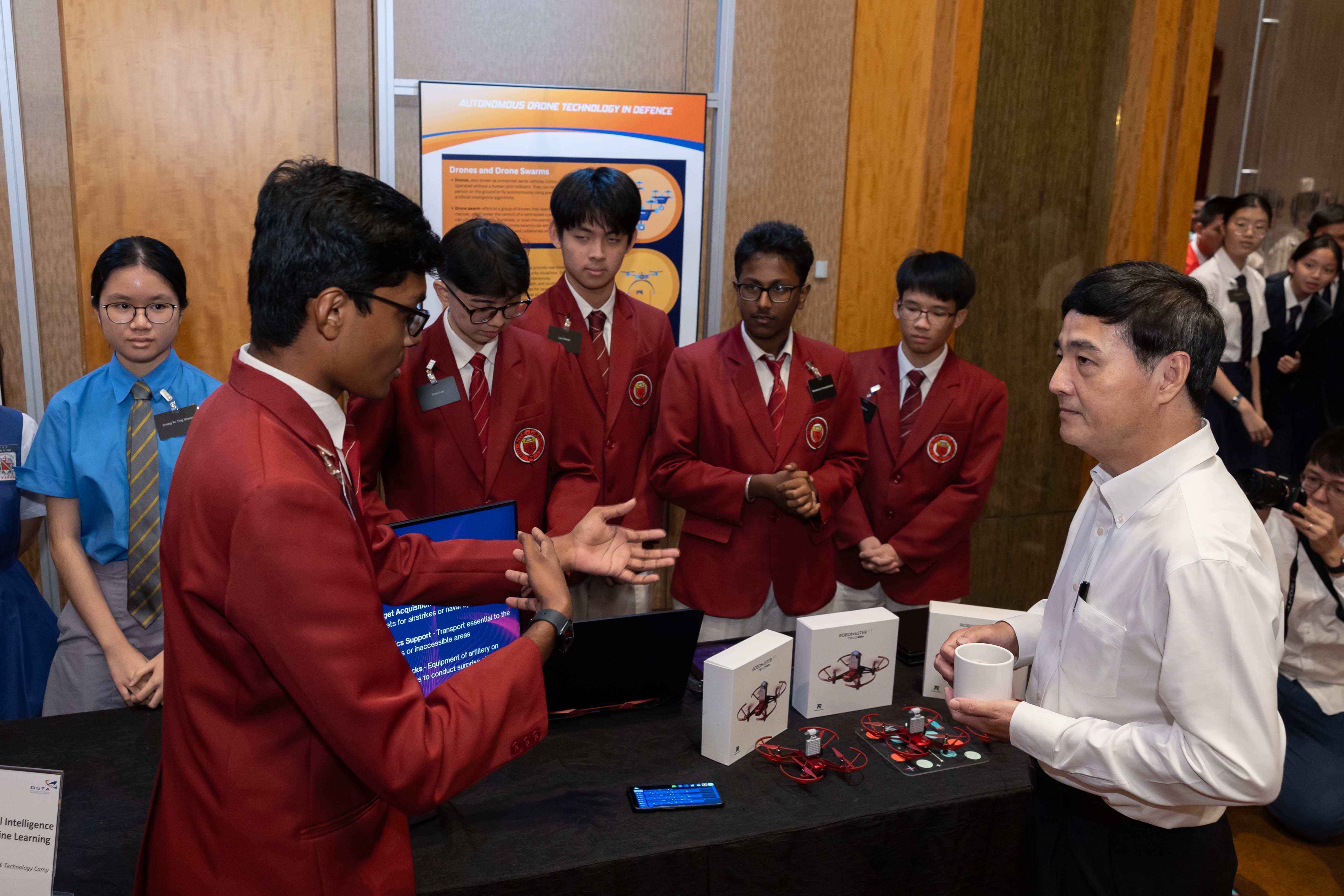 Students in red blazers present a drone technology project to an adult at a exhibition booth with small drones on the table.