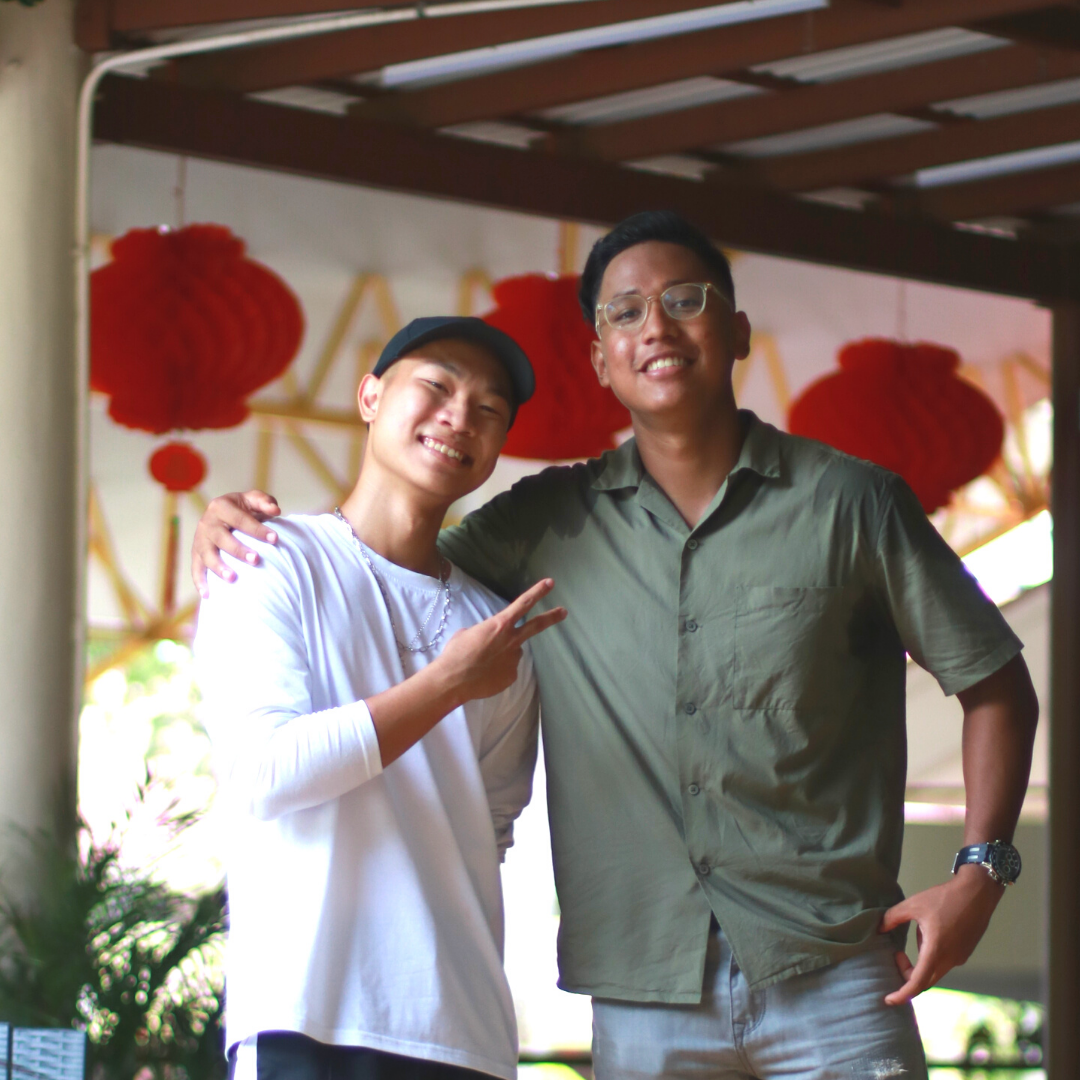 Two smiling young men pose together under a wooden pergola with red Chinese lanterns in the background.