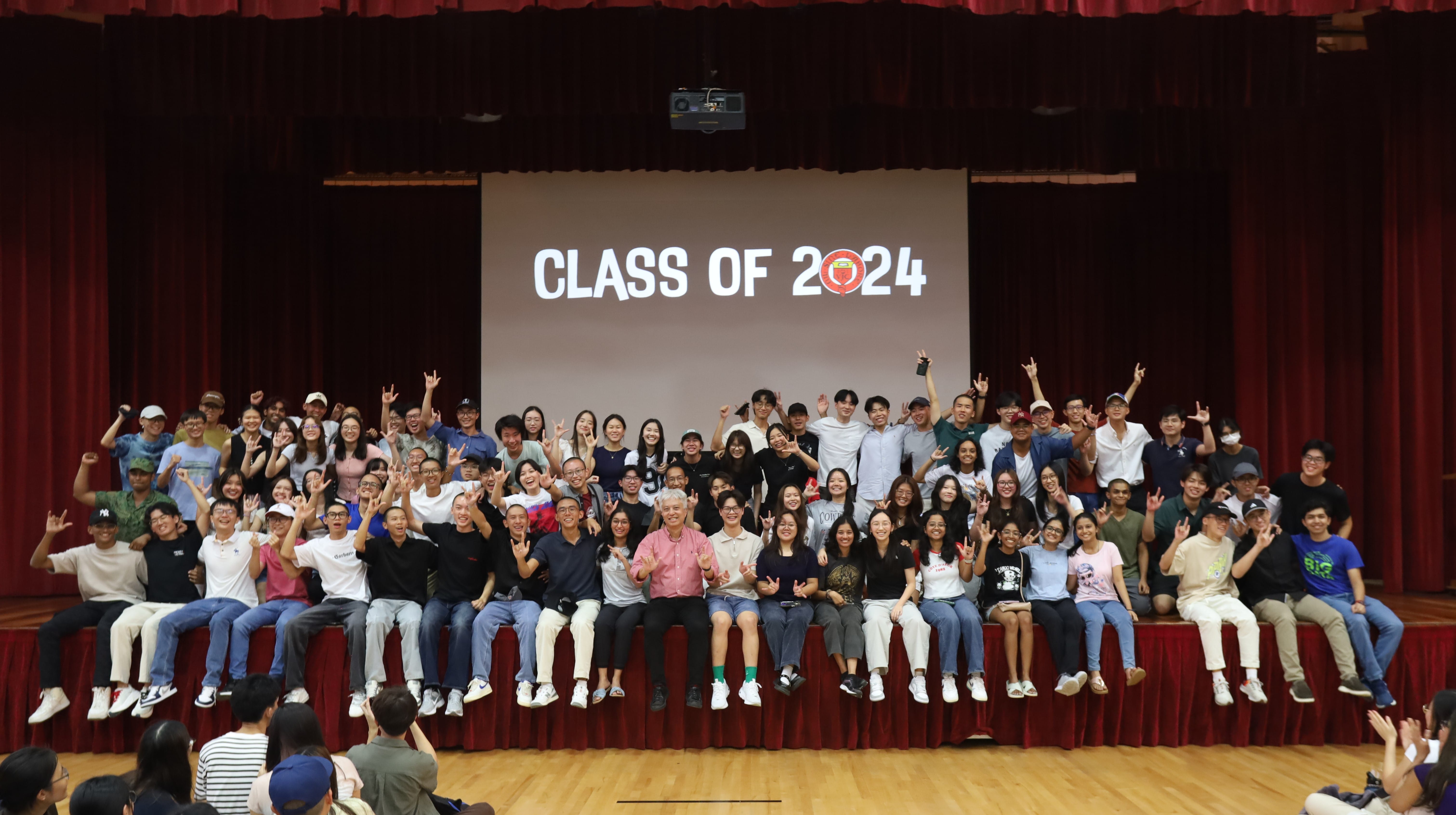 Large group of students posing on a stage in front of a screen displaying CLASS OF 2024 with red curtains.